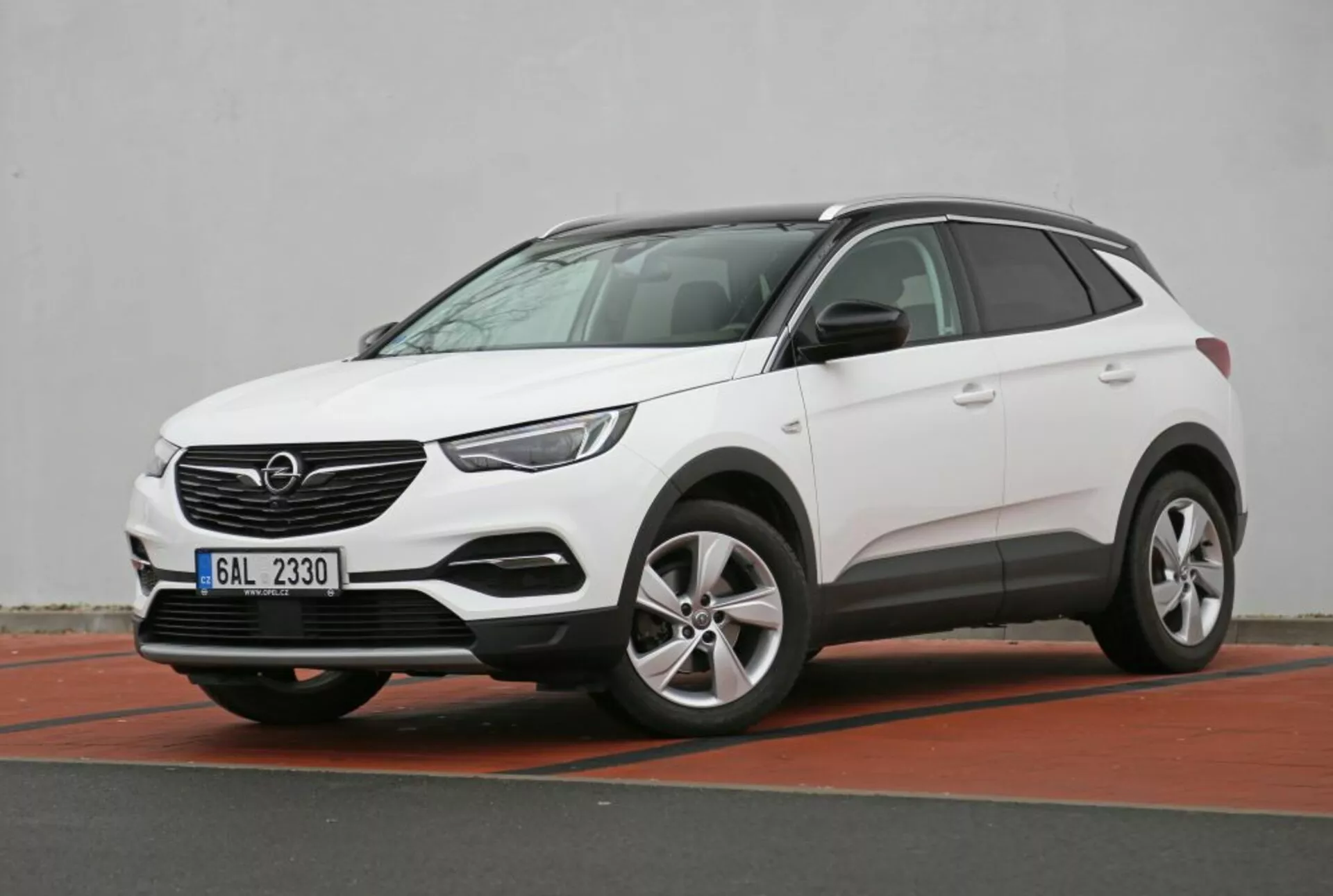 SUV crossover compacto blanco Nissan Qashqai 2014 estacionado en ángulo mostrando sus perfiles frontal y lateral, destacando su altura de conducción desde un ángulo bajo, faros modernos LED, sensores de estacionamiento en el parachoques delantero con una ilusión de placa de deslizamiento y ruedas de aleación, en un entorno de estacionamiento urbano con un bordillo rojo