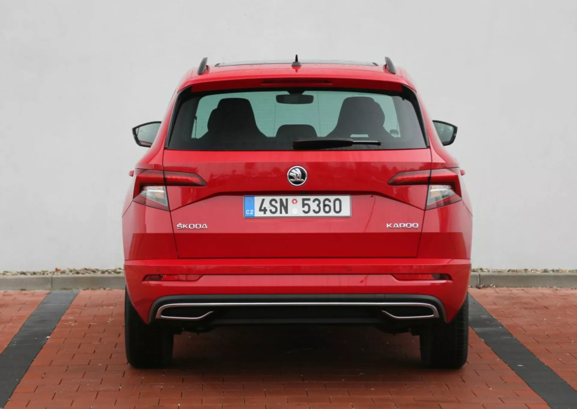 Vista detallada de la parte trasera de un Skoda Kodiaq 2014 en rojo, mostrando el diseño trasero simétrico con el logotipo de Skoda, luces traseras cristalinas, placa de licencia, sensores de aparcamiento en el parachoques, puntas de escape dobles y una antena de aleta de tiburón, contra un fondo neutro con pavimentación estampada.