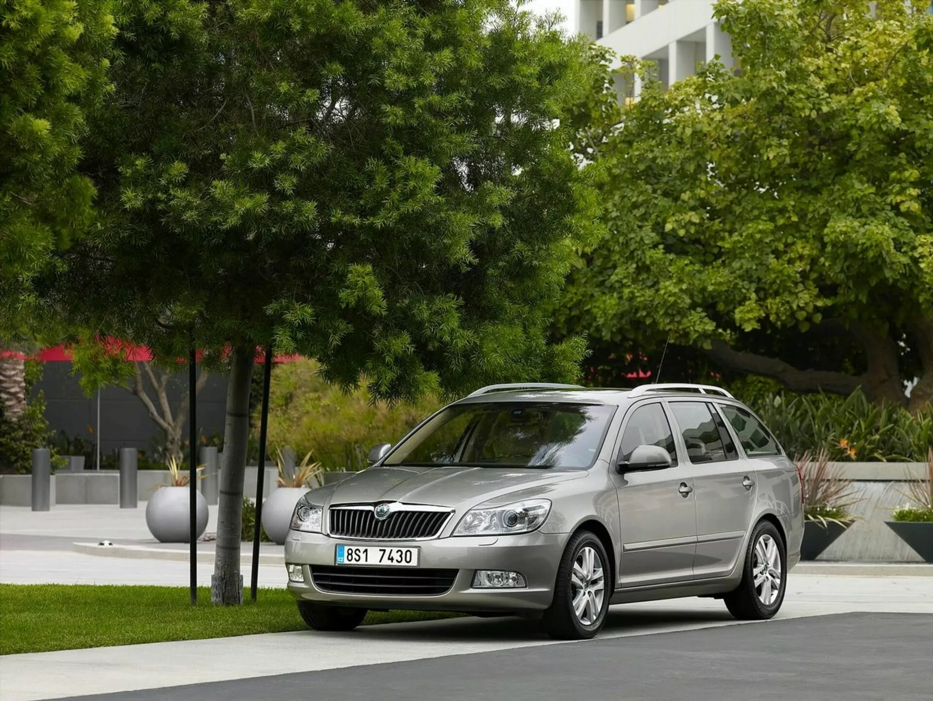 Vagón estacionario Opel Astra de 2011 aparcado en un entorno urbano con vegetación y edificios modernos al fondo, mostrando la parrilla frontal del coche, faros y un perfil lateral elegante incluyendo ruedas de aleación y barras de techo