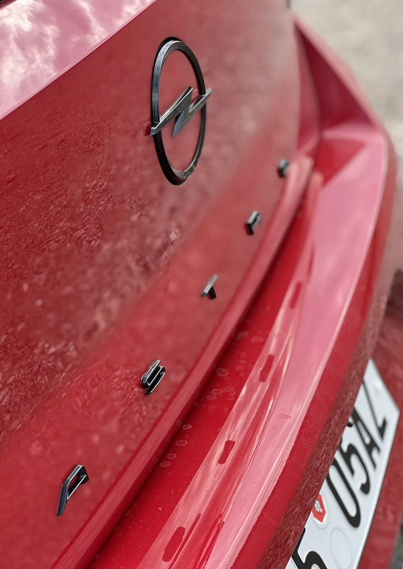 Vista detallada de la sección trasera del Opel Astra 2022 con pintura roja y capa de polvo visible, mostrando el emblema del fabricante y sensores de asistencia para estacionamiento