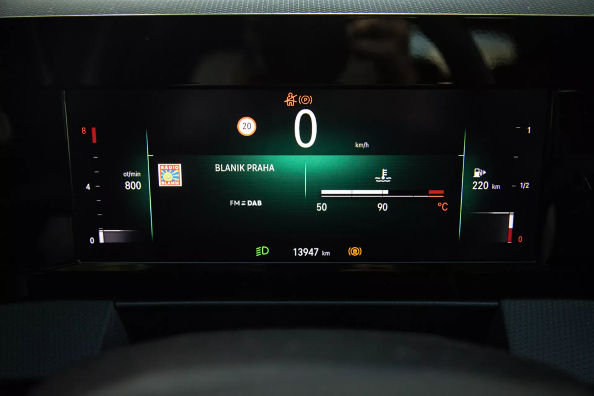 Vista del tablero digital de Opel Astra 2022 destacando el cuadro de instrumentos con RPM, nivel de batería, velocímetro, indicador de combustible, temperatura exterior, estación de radio 'BLANIK PRAHA', y lectura del odómetro desde la perspectiva del asiento del conductor.