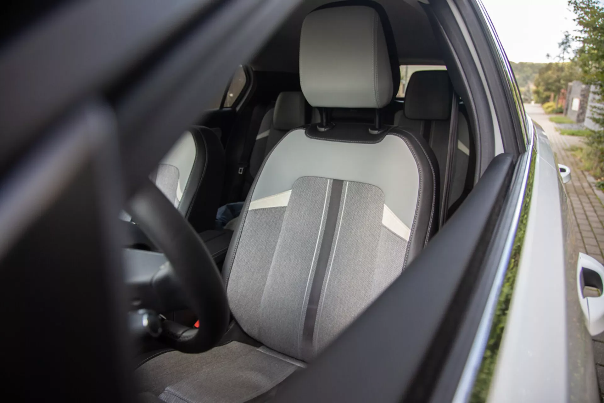 Vista en detalle del interior del lado del conductor del Opel Astra 2022 desde un ángulo lateral externo que muestra el asiento del conductor de tela de dos tonos con reposacabezas integrado, parte del volante con controles, visión parcial del tablero de instrumentos, cinturón de seguridad abrochado, interior de la puerta con posibles controles de ventanas eléctricas y carcasa del espejo lateral del color de la carrocería, con énfasis en características ergonómicas, de seguridad y comodidad.