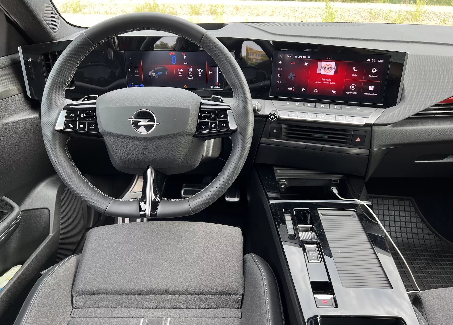 Vista interior del Opel Astra 2022 desde el asiento trasero, mostrando el volante de tres radios con controles integrados, cuadro de instrumentos digital, pantalla táctil de infotenimiento, consola central con palanca de cambios y almacenamiento, y una mezcla de materiales de tela y efecto metálico
