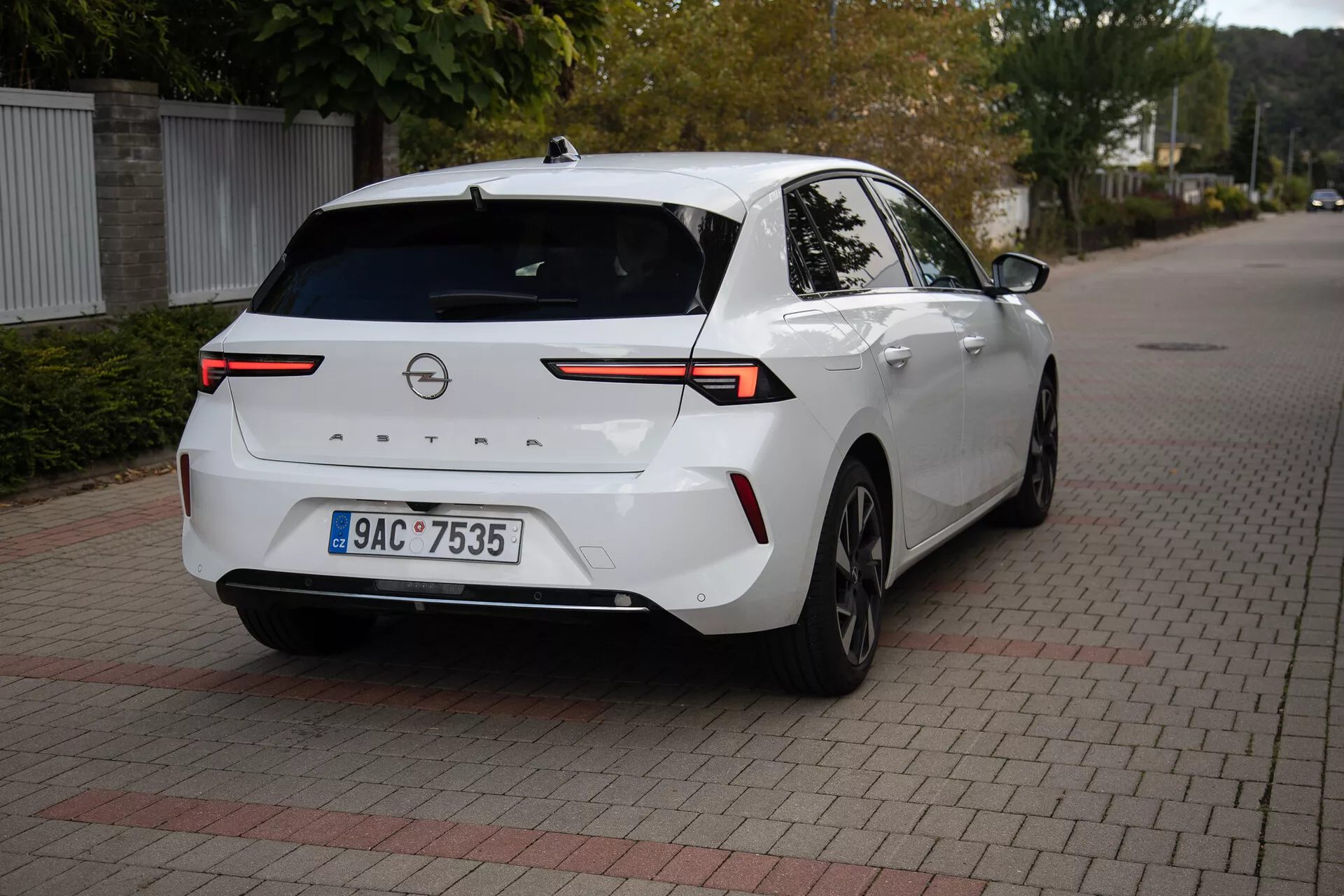 Sedán moderno Opel Astra 2022 en blanco, aparcado sobre superficie de ladrillos entrelazados, mostrando ángulo trasero de tres cuartos con enfoque en luces traseras LED de alta tecnología, cámara trasera, sensores de aparcamiento, perfil lateral parcial incluyendo la rueda y puerta trasera derecha, conectadas por una barra de luz horizontal a través de la tapa del maletero, con antena de aleta de tiburón para una conectividad mejorada, adecuado para ilustrar el diseño trasero del vehículo y la tecnología automotriz moderna