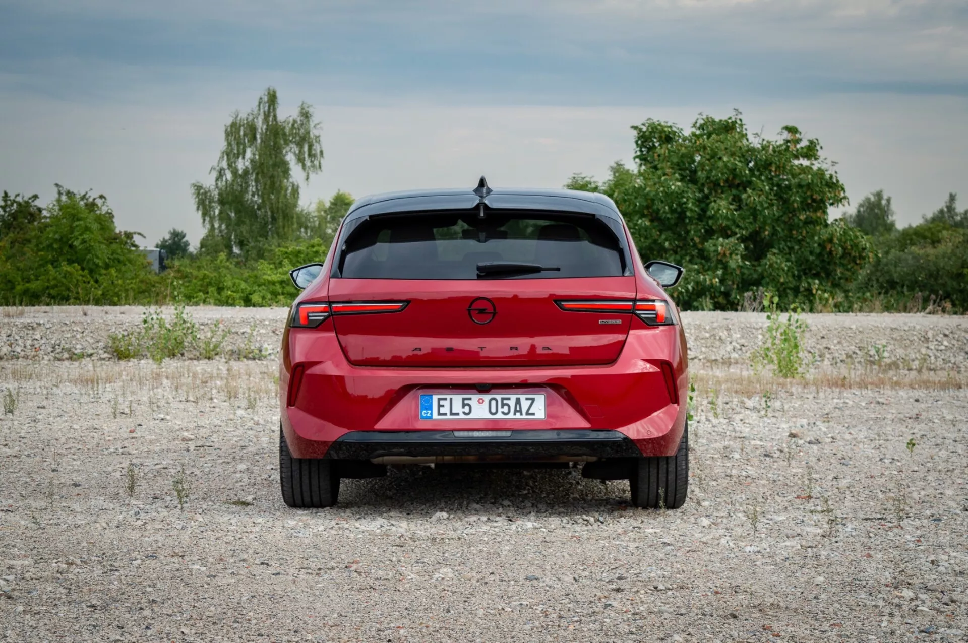 Opel Astra 2022 fotografiado desde la parte trasera sobre una superficie de grava mostrando las luces traseras con una barra de luz conectando, sensores de aparcamiento y ausencia de puertos de escape indicando un posible modelo eléctrico o híbrido, contra un telón de fondo de vegetación y cielo ligeramente nublado.