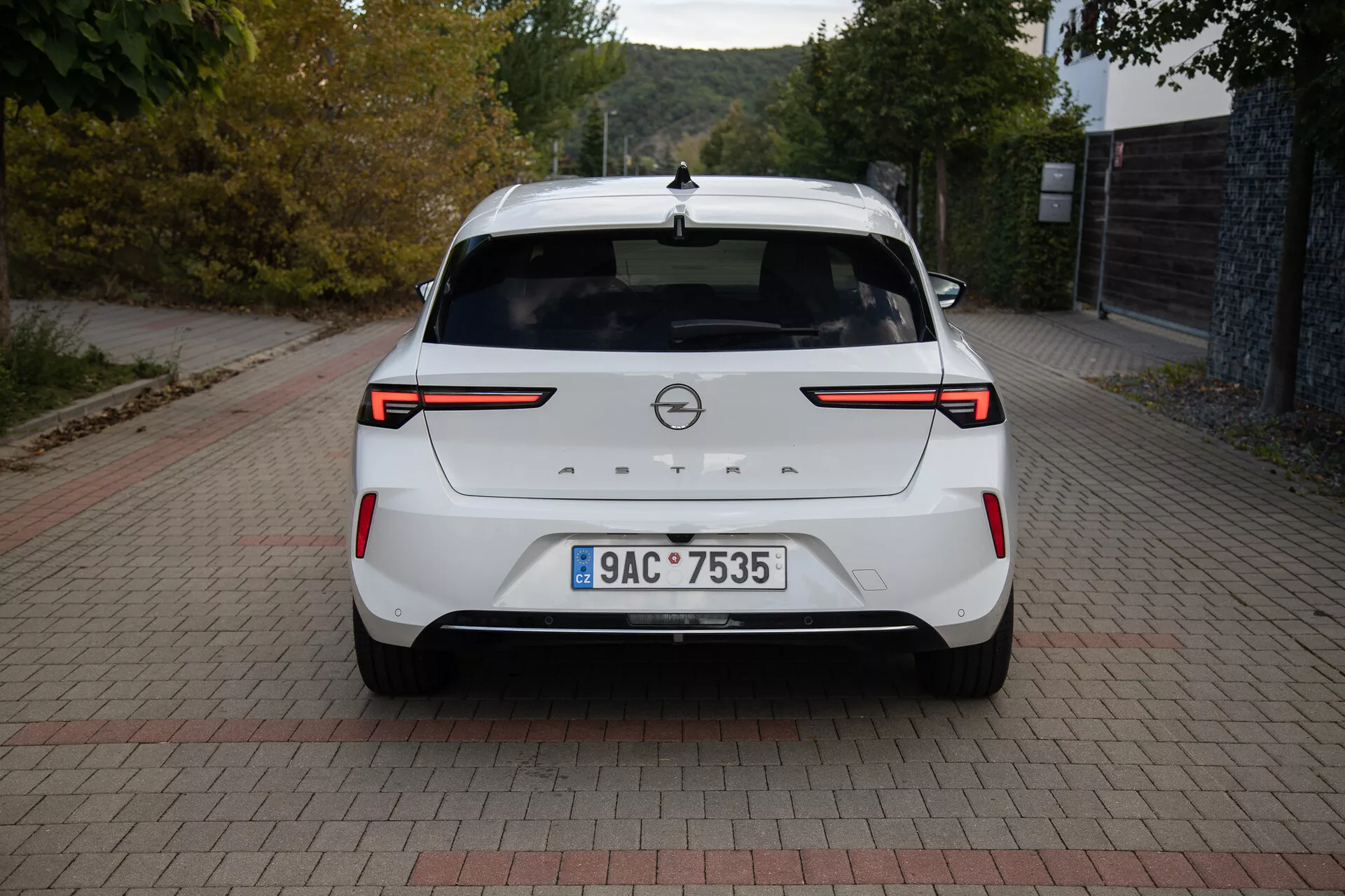 Vista trasera de un Opel Astra 2022 blanco mostrando luces traseras LED, sensores/cámara de estacionamiento trasero, emblema de la marca, placa de matrícula, alerón trasero, luz de freno de montaje alto, antena de aleta de tiburón y un acabado de pintura limpio, estacionado en una superficie con patrón de ladrillos a la luz del día.