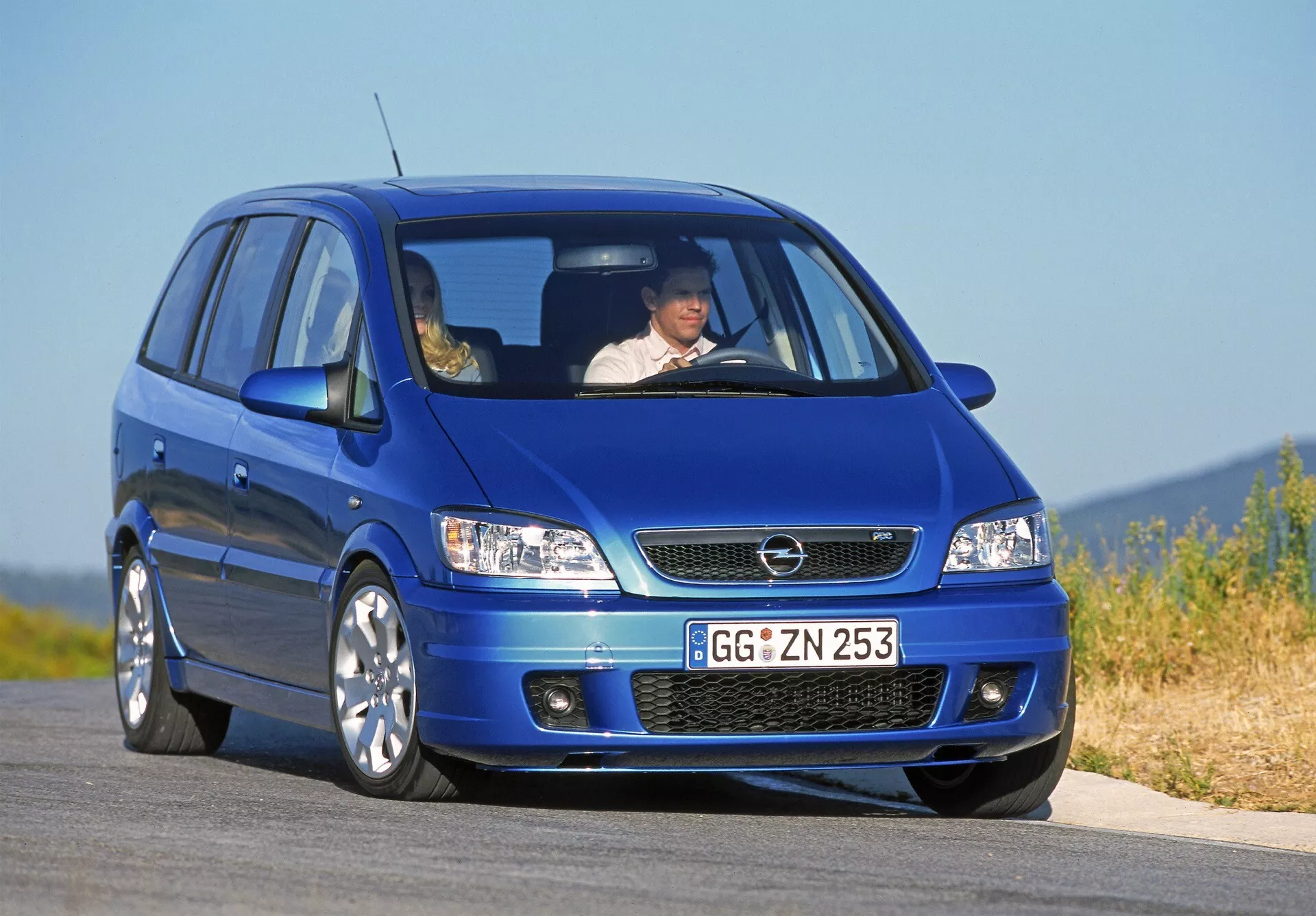 Imagen de un Opel Zafira (A-facelift) compacto MPV azul de 2002 desde una vista baja tres cuartos delantera, destacando la fascia delantera del coche con visibilidad clara de la parrilla, faros halógenos con señales de giro integradas, parachoques delantero con toma de aire y ventilaciones laterales auxiliares, capó aerodinámico con contornos sutiles, grandes llantas de aleación multirradio y un voladizo delantero corto que sugiere una disposición de tracción delantera. A través del parabrisas delantero se puede ver a un conductor con el cinturón de seguridad puesto, subrayando las características de seguridad vial.