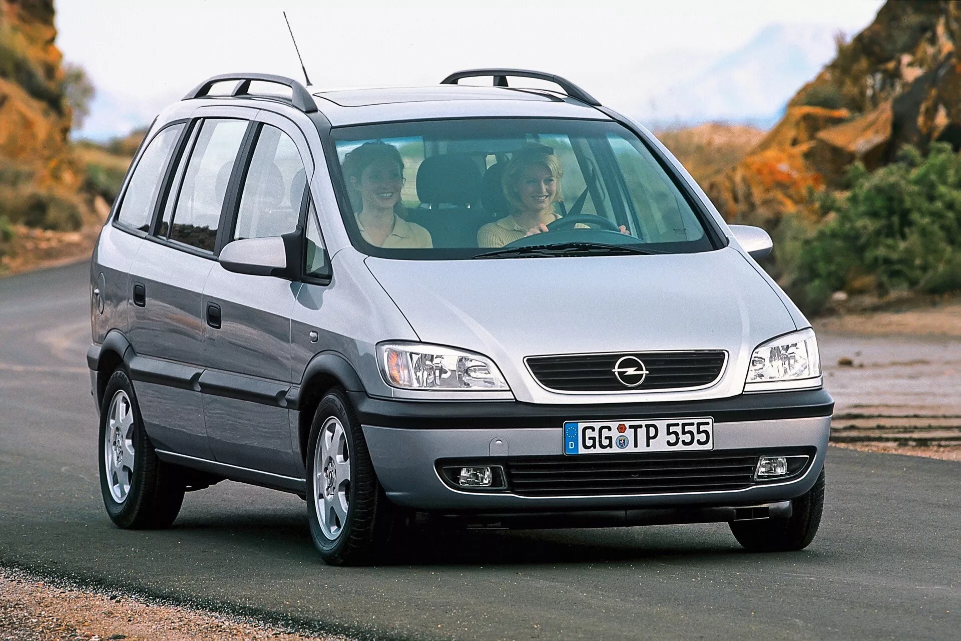 Fotografía de un Opel Zafira de 1999, un MPV compacto, tomada desde una perspectiva baja de tres cuartos frontales. La imagen resalta la parrilla del coche con una insignia central, grandes faros probablemente con fuentes de luz halógenas, capó liso, parachoques delantero con luces antiniebla, llantas de aleación y el lado del pasajero del vehículo con una antena en la parte trasera del techo. El vehículo está en una carretera pavimentada con un paisaje natural de fondo. A través del parabrisas delantero, se pueden ver dos ocupantes, reflejando el diseño interior y la amplitud del coche.