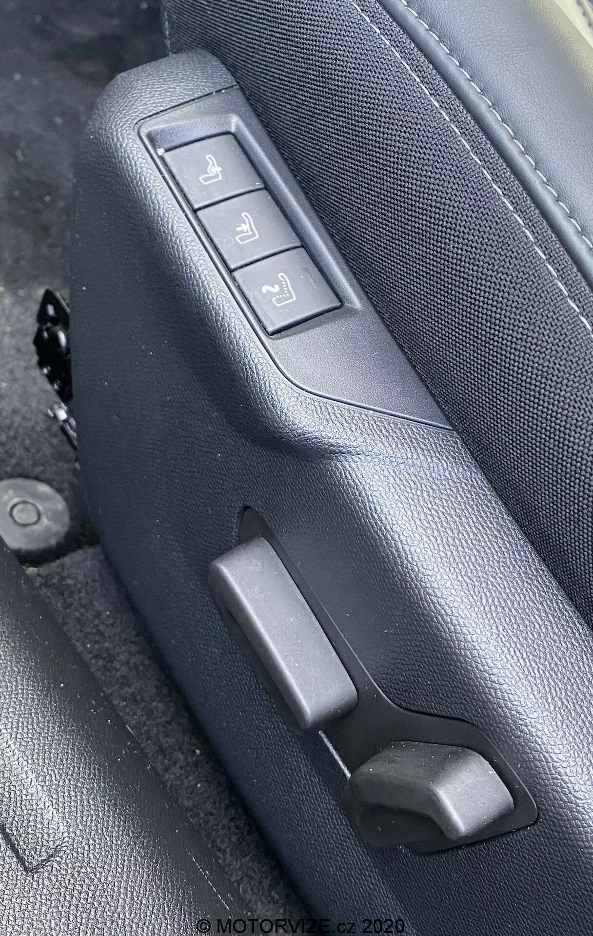 Imagen de primer plano de los controles del asiento interior del Peugeot 2008 1.2 PureTech 100 Active (2020), destacando el lado de un asiento de cuero negro o gris oscuro con costuras detalladas, visto desde un ángulo oblicuo ligeramente elevado. El asiento presenta tres botones de ajuste electrónico y una palanca manual. Los botones gestionan el movimiento hacia adelante/atrás, la inclinación del respaldo y la altura total del asiento, montados en un panel texturizado a juego. La marca de agua dice 'MOTORVIZE.cz 2020'.