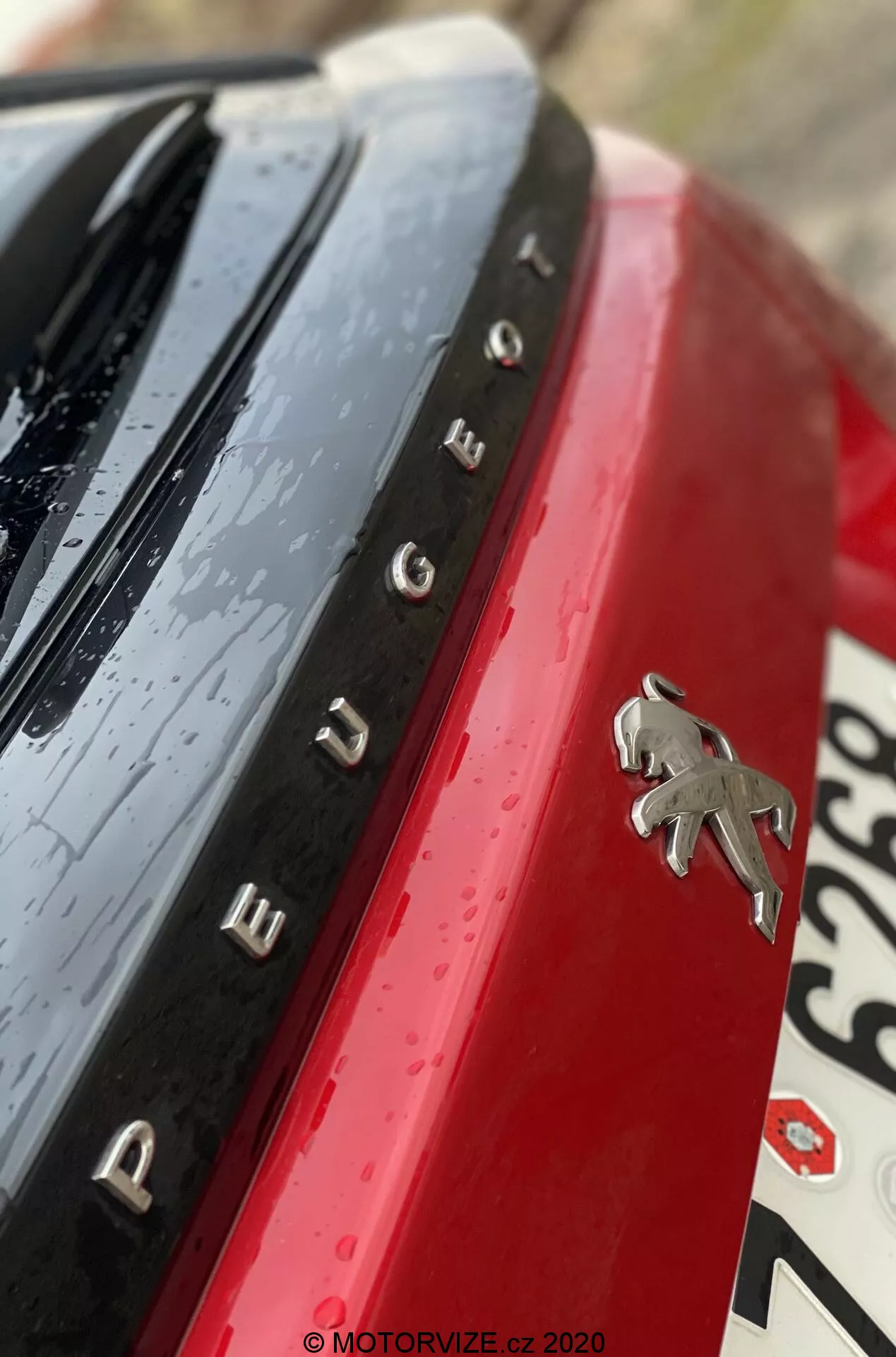 Detalle en primer plano del emblema trasero y la designación del modelo Peugeot 2008 2020 (1.2 PureTech 100 Active). La toma está en un ángulo diagonal poco ortodoxo, capturando el logo de Peugeot en cromo contra un panel de carrocería rojo vibrante. Letras cromadas espaciadas a la izquierda del logo deletrean 'PEUGEOT'. Una superficie negra brillante con gotas de agua aparece encima del nombre, indicando lluvia reciente o un lavado de automóvil. La sección de la matrícula borrosa es visible abajo a la derecha. La poca profundidad de campo enfoca el emblema y el nombre, mientras que otros elementos están desenfocados para un toque artístico.