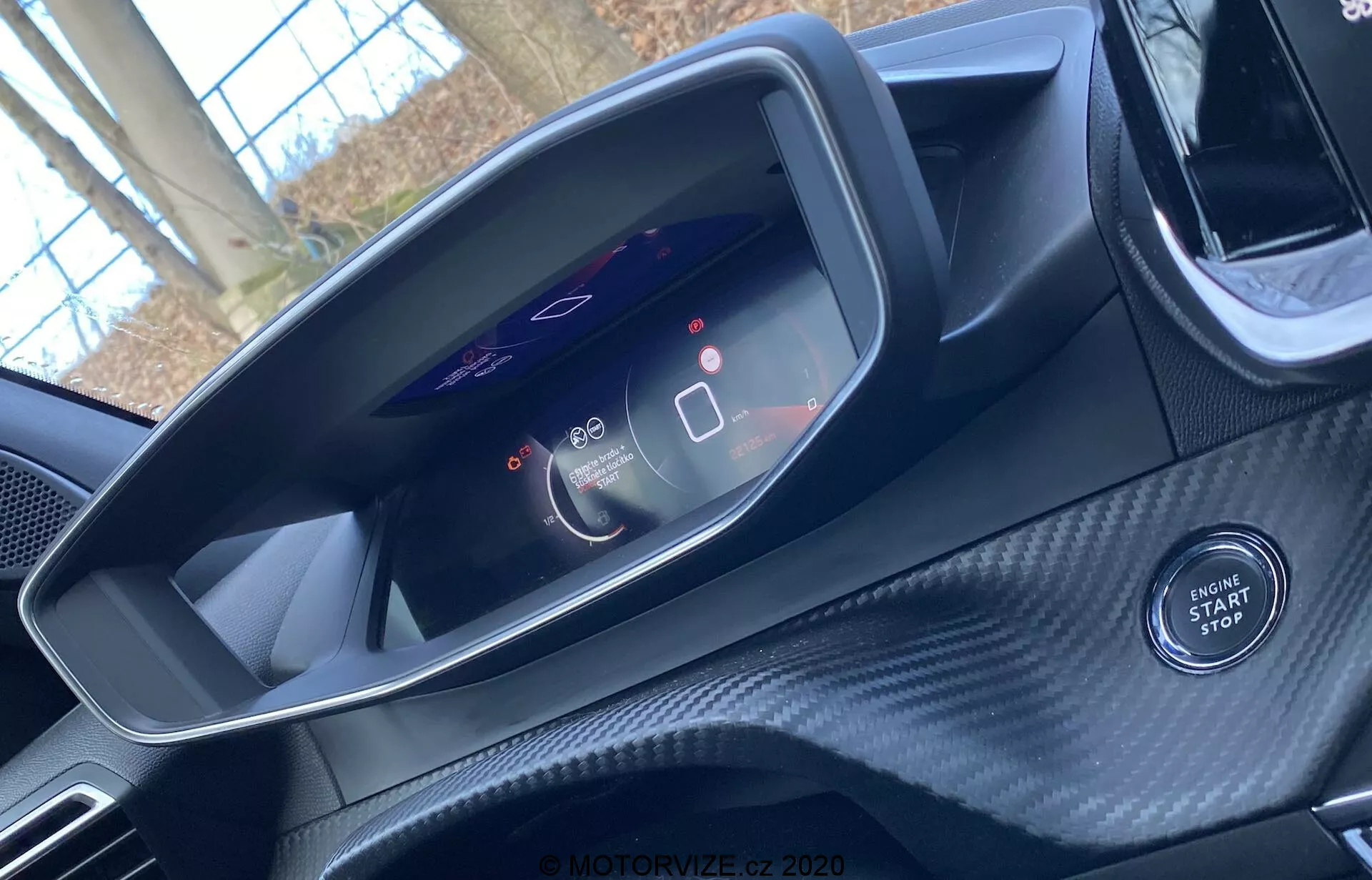 Vista interior de un Peugeot 2008 2020 desde la perspectiva del asiento del conductor. La imagen se centra en el moderno panel de instrumentos digital que muestra un velocímetro a 0 y un medidor de uso de energía. También muestra iconos de sistemas avanzados de asistencia al conductor. El botón de arranque/parada del motor es visible debajo del panel, lo que indica el encendido sin llave del coche. El área circundante tiene una textura similar a la fibra de carbono, lo que sugiere un interior deportivo o de alta gama. La parte superior del volante es apenas visible.