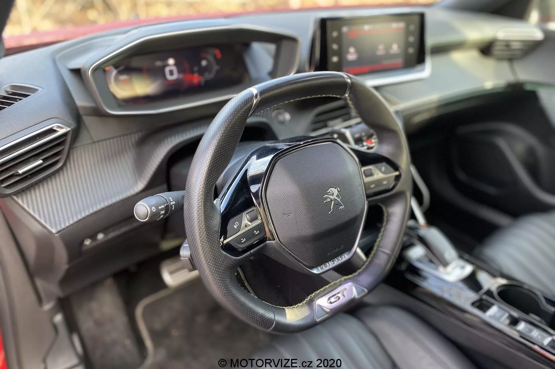 Interior del Peugeot 2008 1.2 PureTech 100 Active de 2020 desde la perspectiva del pasajero delantero enfocado en la cabina del conductor. La imagen muestra un volante de fondo plano con múltiples botones de control y el logo del león de Peugeot, un cuadro de instrumentos digital, sistema de infoentretenimiento con pantalla táctil, consola central de transmisión automática, botón de encendido/apagado, salidas de aire con acentos cromados y una mezcla de materiales suaves al tacto, incluyendo cuero y texturas similares a la fibra de carbono.