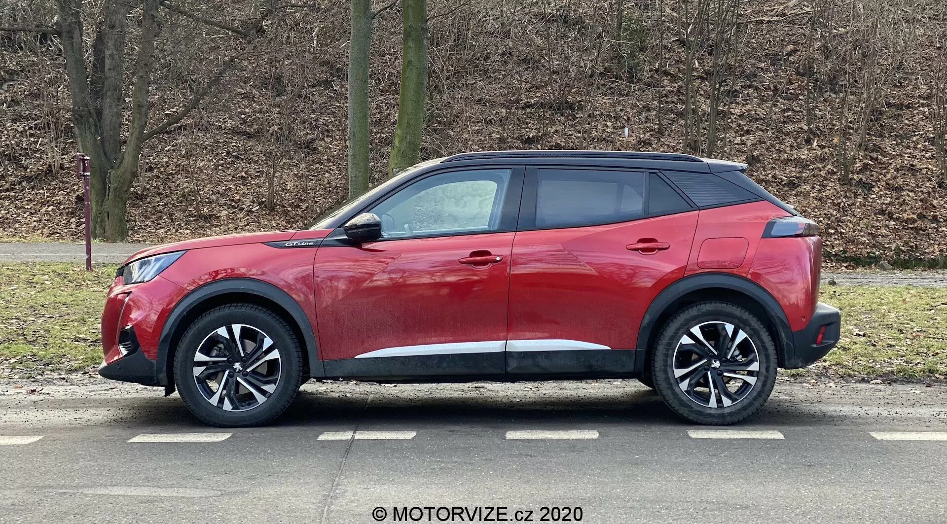 Vehículo utilitario deportivo (SUV) Peugeot 2008 2020 1.2 PureTech 100 Active rojo con diseño moderno y elegante, línea de techo inclinada, línea de cintura alta con ventanas que se estrechan, manijas de puertas aerodinámicas, espejos laterales con intermitentes integrados, grandes llantas de aleación de múltiples radios, neumáticos de perfil bajo, molduras de plástico negro para una apariencia robusta, acabado de pintura roja brillante y ventanas traseras tintadas, captado en vista de perfil estacionado en un entorno al aire libre con árboles sin hojas y fondo de césped.