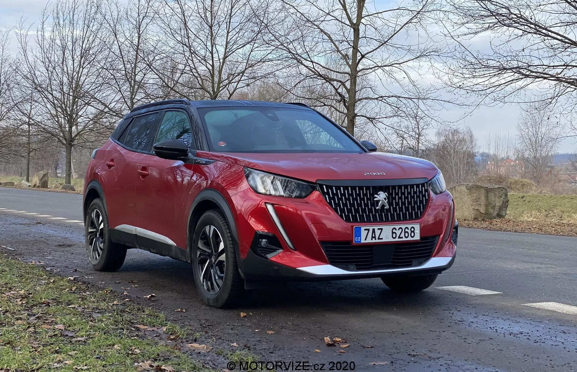SUV rojo Peugeot 2008 2020 (Peugeot-2008-1-2-Puretech-100-Active) desde la vista tres cuartos frontal, destacando la rejilla delantera con un emblema de marca prominente, faros LED elegantes, sensores de estacionamiento delanteros y luces antiniebla. La vista lateral revela ruedas de aleación multirradio, altura de manejo mayor, líneas de carrocería dinámicas, señales de giro integradas en los espejos laterales y revestimiento de plástico negro. Posicionado en un ángulo leve al costado de una carretera con un paisaje rural natural en el fondo, indicando finales de otoño o principios de primavera. Ángulo de cámara bajo para una vista poderosa e imponente.
