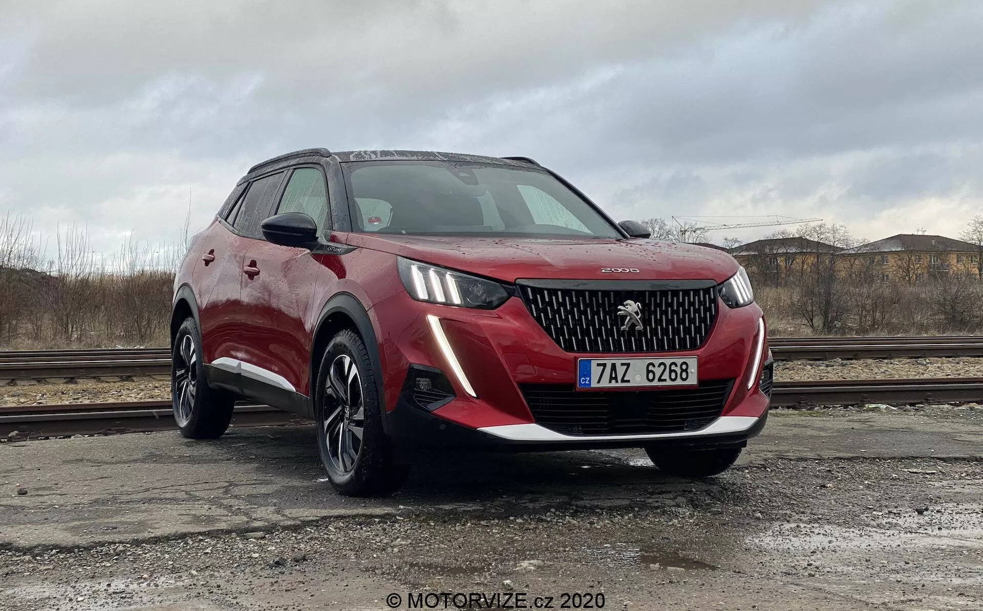 Vista de tres cuartos delanteros de un Peugeot 2008 1.2 PureTech 100 Active 2020 rojo, mostrando su rejilla detallada, faros LED, líneas de contorno nítidas en el capó y elegantes luces diurnas. La imagen captura el perfil lateral del lado del conductor, incluyendo el espejo retrovisor y la línea de ventanas, destacando el diseño elegante y las características tecnológicas avanzadas del vehículo. El coche está posicionado probablemente sobre una superficie de concreto, con reflejos y texturas claramente visibles en la carrocería, presentando una perspectiva equilibrada adecuada para fines de marketing.