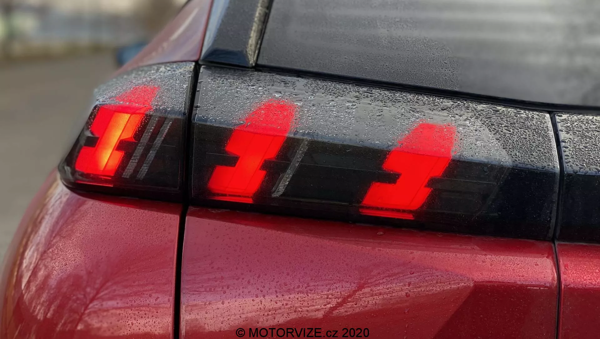 Primer plano de la sección trasera de un Peugeot 2008 2020 rojo (PureTech 100 Active) con enfoque en el conjunto de luces traseras que muestran las luces de freno iluminadas. La fotografía, tomada desde la esquina trasera ligeramente hacia el lado, resalta tres elementos de luz LED verticales distintos y gotas de agua en la superficie, destacando el diseño moderno de la tecnología de iluminación automotriz.