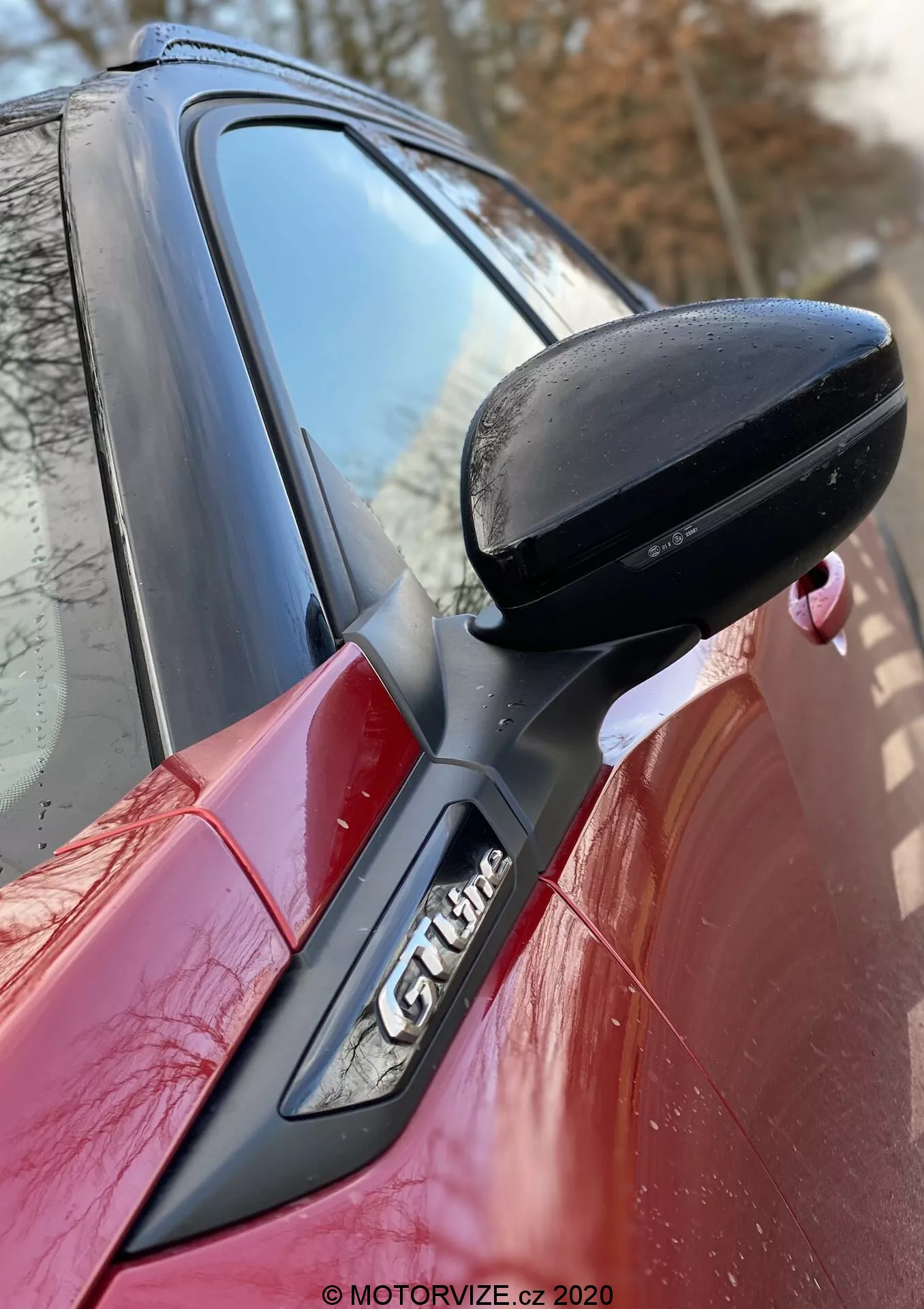Imagen en primer plano del lado de un Peugeot 2008 2020 rojo, enfocando el espejo retrovisor lateral del conductor negro, montado en la puerta con soporte negro. El pilar A con el emblema 'GT Line' es parcialmente visible, mostrando las líneas de diseño aerodinámico del coche y las características del acabado deportivo. Marca de agua de MOTORVIZE.cz 2020.