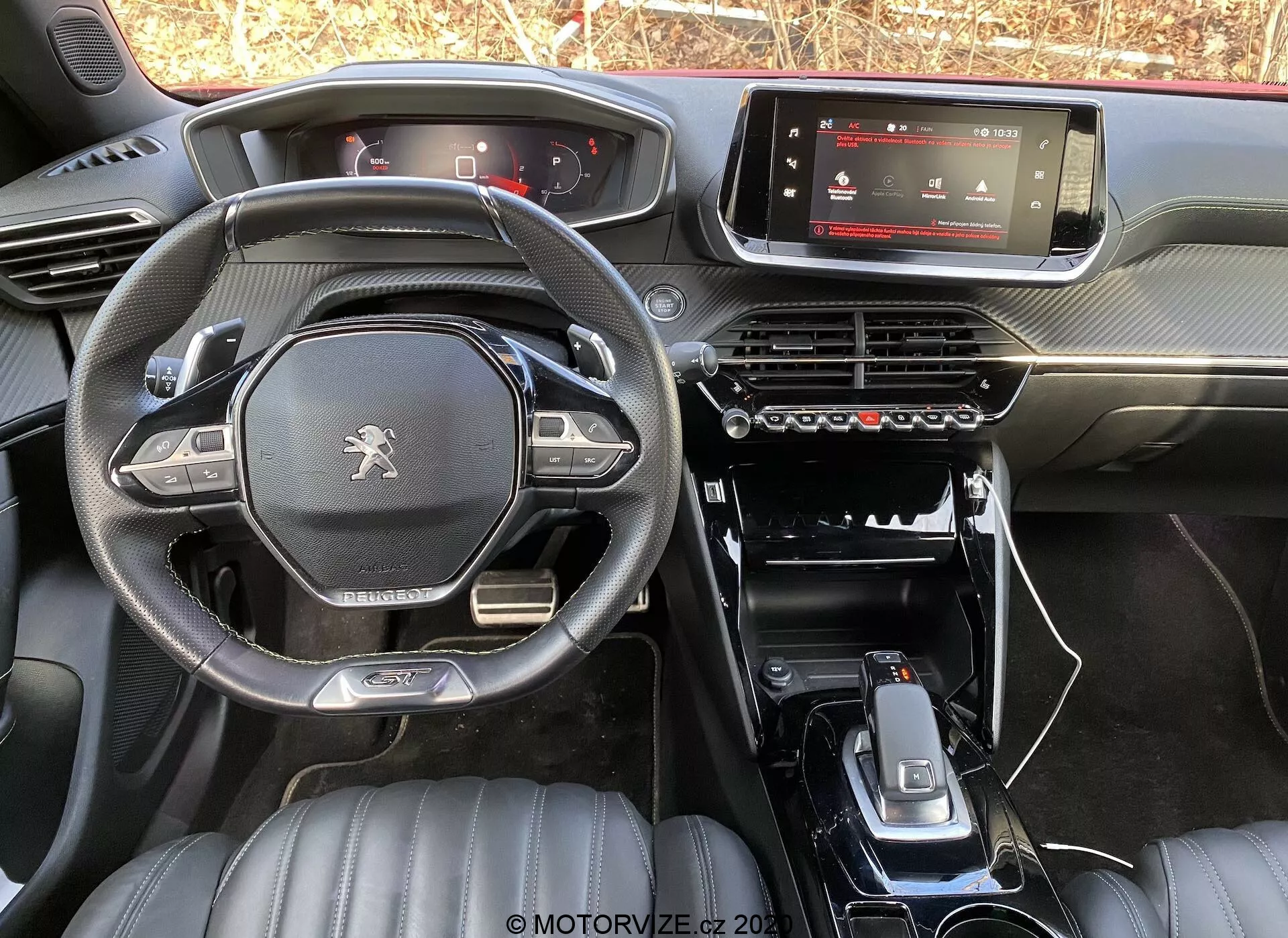Vista interior del Peugeot 2008 1.2 PureTech 100 Active 2020 desde el asiento trasero, que muestra el salpicadero, la pantalla digital del sistema de infoentretenimiento, el panel de control de clima, la consola central minimalista con palanca de cambios, portavasos y botones de control adicionales. El volante de fondo plano tiene el logotipo de Peugeot y controles multifuncionales. Los asientos delanteros de cuero con patrones acolchados sugieren un nivel de acabado superior. La iluminación natural resalta el diseño y los materiales del modelo de fábrica.