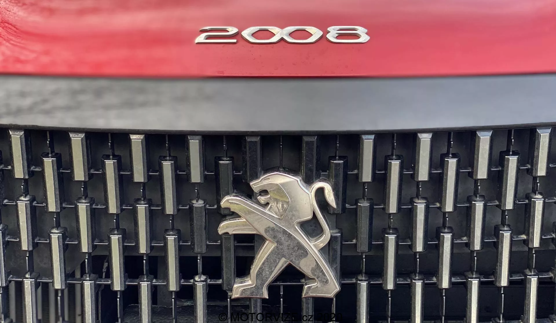 Vista de primer plano de la parrilla delantera de un Peugeot 2008 1.2 PureTech 100 Active 2020, que muestra el diseño distintivo con un capó rojo vibrante, una compleja array de listones verticales y horizontales negros, y un emblema de león plateado pulido en el centro. La foto se enfoca en los elementos estéticos y de marca, proporcionando una visualización clara de los detalles de la parrilla.