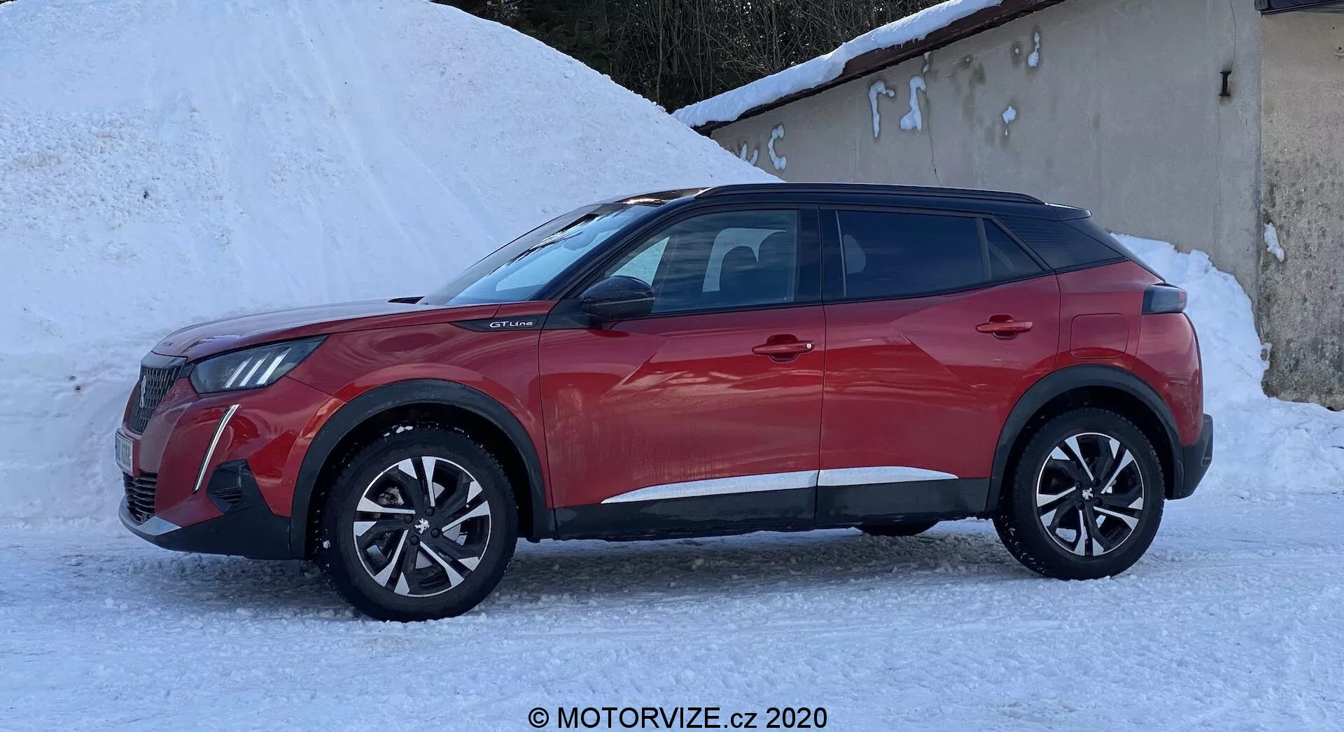 Toma de perfil lateral de un Peugeot 2008 2020 1.2 PureTech 100 Active SUV crossover de cuatro puertas en rojo. El vehículo está en un entorno nevado, lo que resalta sus posibles capacidades en clima frío. Cuenta con ruedas de aleación de diseño multirradio, molduras de plástico negro en la parte inferior de las puertas y los arcos de las ruedas, y ventanas elegantes. El diseño de los faros y la parrilla se vislumbran desde el frente, mostrando una apariencia moderna con posibles faros LED. Los espejos laterales incluyen intermitentes integrados. La línea del techo inclinada y el sistema de rieles para techo sugieren practicidad y utilidad para un estilo de vida activo.