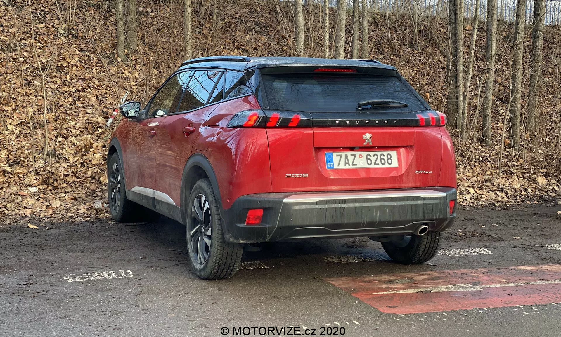 Vista en ángulo trasero de un SUV Peugeot 2008 1.2 PureTech 100 Active rojo del 2020 estacionado en una superficie pavimentada, con un fondo de árboles sin hojas que sugiere finales de otoño o invierno. Capturado desde una perspectiva trasera de tres cuartos, enfatizando el lado derecho del vehículo, la imagen muestra elementos de diseño como las luces traseras, la ventana trasera, el parachoques trasero de dos tonos con sensores de estacionamiento, el alerón del techo, las franjas verticales de las luces de freno, las ruedas de aleación parciales y las barras del techo. El escape oculto sugiere un sistema de propulsión potencialmente híbrido o eléctrico. El vehículo cubre la mayor parte del cuadro, indicando la proximidad del fotógrafo.