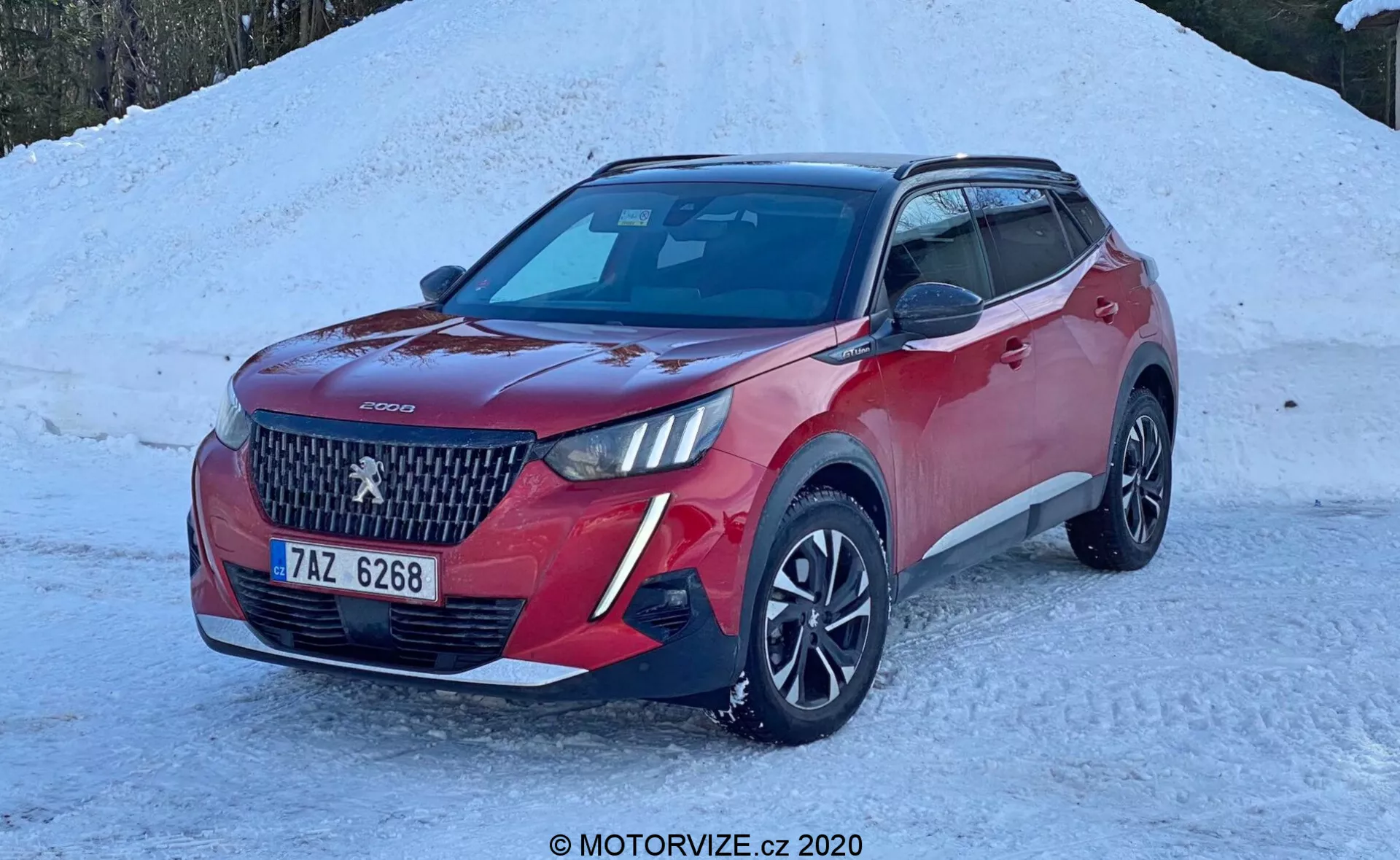 Imagen de un SUV rojo, concretamente un Peugeot 2008 2020 (peugeot-2008-1-2-puretech-100-active), aparcado en un terreno cubierto de nieve. El vehículo moderno se muestra en un ángulo de tres cuartos desde el frente, destacando su prominente parrilla con un gran logotipo central y diseño de patrón de diamante. Los faros LED elegantes, la pintura de carrocería bicolor con tapas de espejo negras y techo, llantas de aleación de diseño multirrayo y las luces antiniebla o adicionales en el parachoques delantero son visibles. El coche cuenta con sensores de aparcamiento delanteros, un módulo de cámara o sensor en el parabrisas para la tecnología de asistencia al conductor y una antena en forma de aleta de tiburón en el techo, sugiriendo funcionalidad GPS o radio satelital. La escena indica un entorno invernal con una luz natural brillante que resalta los detalles del coche.
