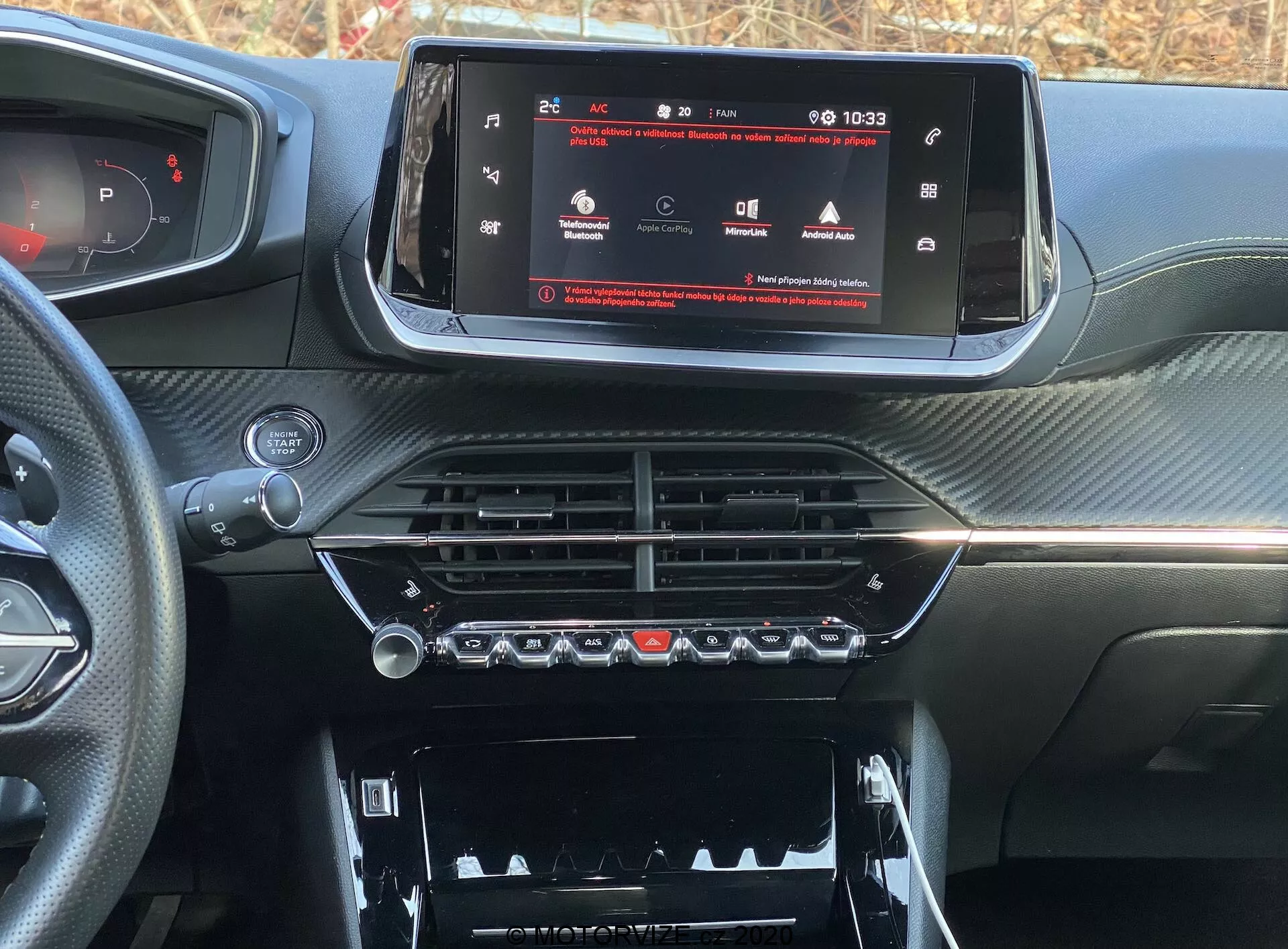 Vista de cerca del interior del salpicadero de un coche moderno, específicamente el Peugeot 2008 2020 (peugeot-2008-1-2-puretech-100-active). La imagen está tomada desde el asiento del conductor o del pasajero delantero, enfocándose en la consola central y el sistema de infoentretenimiento. Una pantalla táctil panorámica muestra un menú de configuración del vehículo, indicando tecnología avanzada. Debajo hay ventilaciones cromadas y controles climáticos con botones físicos y diales giratorios. Más abajo hay un compartimento con una cubierta visible. El salpicadero tiene un acabado texturizado similar a la fibra de carbono, dando una apariencia deportiva y de lujo.
