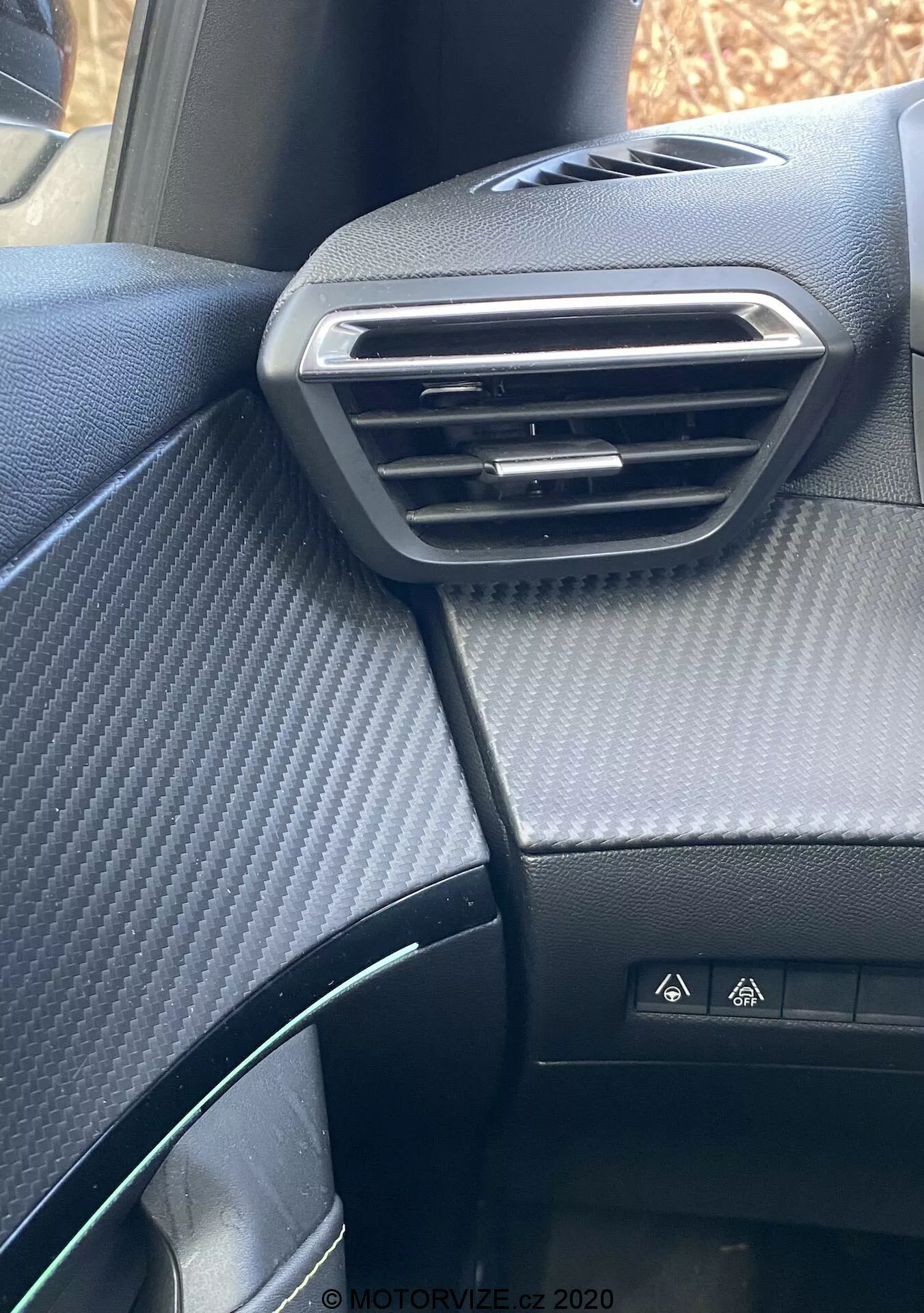 Vista de cerca del salpicadero del lado del pasajero del interior del Peugeot 2008 2020, destacando el ventilador ajustable con acento plateado, la parte superior del salpicadero con textura de fibra de carbono, el botón de luz de emergencia, la parte parcial del volante y el asiento negro con patrón de puntos.