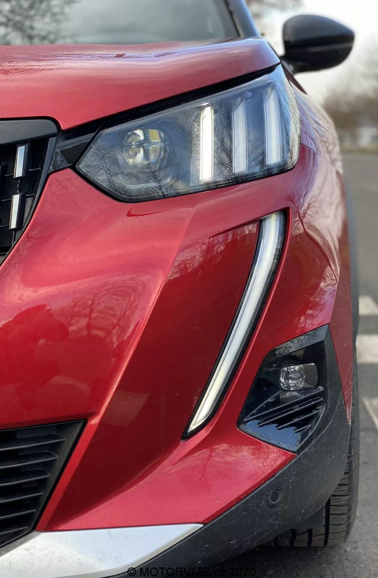 Vista frontal angular en primer plano de un Peugeot 2008 1.2 PureTech 100 Active rojo de 2020, que muestra el cuarto delantero del lado del conductor. Faro prominente con haz proyector e integrados LED DRL, parrilla de listones horizontales y ensamblaje de luz secundaria parcialmente visible dentro de un recorte angular del parachoques. Pintura brillante con acabado metálico, línea de carrocería afilada, contorno elevado del capó, espejo retrovisor negro con posibles señales integradas o cámara de punto ciego, y recorte en la esquina inferior del parachoques que sugiere sistemas avanzados de asistencia al conductor.