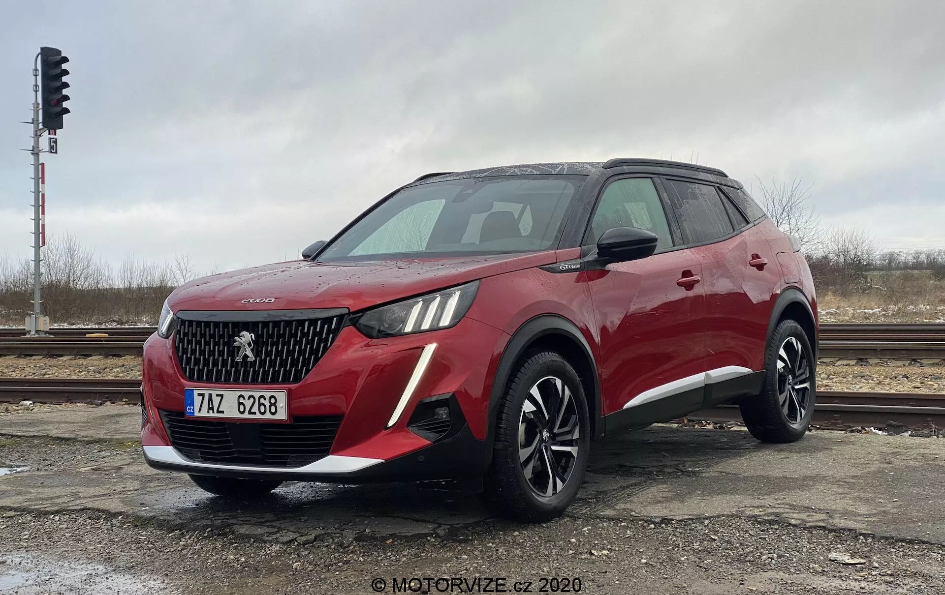 Vista frontal tres cuartos de un moderno SUV crossover rojo Peugeot 2008 1.2 PureTech 100 Active. La toma desde un ángulo bajo destaca la altura y la postura del vehículo. El frente muestra una parrilla prominente con patrón de malla en forma de diamante, un emblema que probablemente representa la insignia del fabricante y faros distintivos que incorporan luces diurnas LED. El parachoques delantero contiene luces antiniebla o entradas de aire. El perfil lateral revela líneas aerodinámicas, grandes llantas de aleación de múltiples radios, rieles de techo y espejos laterales con luces intermitentes integradas. La imagen captura el diseño y las características tecnológicas del SUV en un día nublado, estacionado cerca de vías férreas.