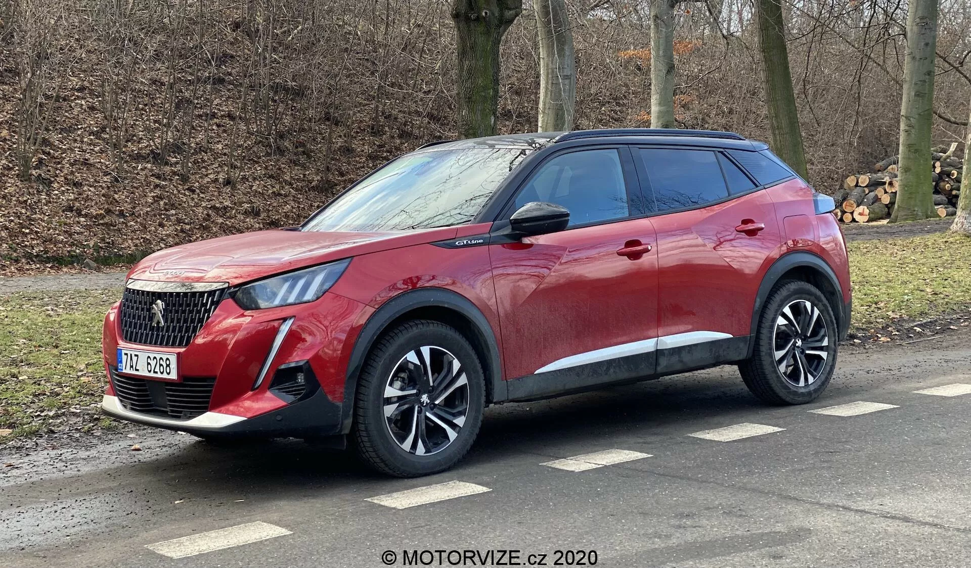 La imagen muestra un Peugeot 2008 1.2 PureTech 100 Active del 2020, un SUV moderno estacionado en ángulo en la carretera. Esta perspectiva de tres cuartos resalta el frente y el lado del pasajero del vehículo, revelando su elegante pintura roja, líneas dinámicas del cuerpo, parrilla distintiva, faros LED afilados y llantas de aleación de múltiples radios. Los arcos pronunciados de las ruedas, la línea de cintura ascendente y los detalles cromados debajo de las ventanas tintadas mejoran su apariencia premium. La línea del techo se inclina suavemente con rieles de techo visibles, lo que sugiere versatilidad deportiva. Una parte trasera parcialmente visible sugiere luces traseras LED envolventes y un spoiler integrado.