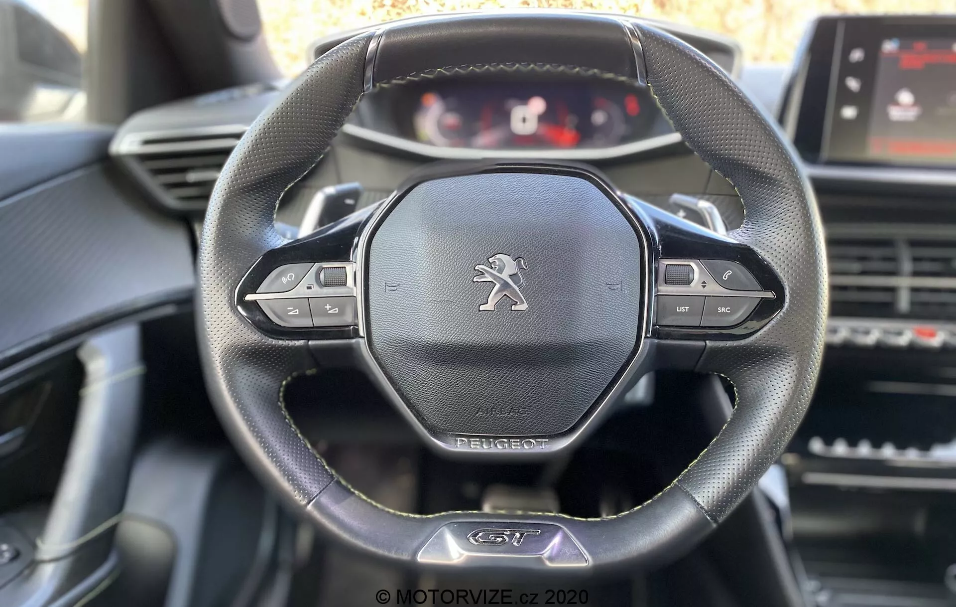 Vista interior del Peugeot 2008 2020 enfocado en el volante y el tablero desde la perspectiva del conductor. Incluye características destacadas como el volante de cuero de tres radios con botones multifuncionales, el emblema del león de Peugeot y el panel de instrumentos digital personalizable. Elementos visibles incluyen pantalla táctil de infoentretenimiento, ventilaciones y controles de la consola central con una suave iluminación diurna destacando los detalles del volante.