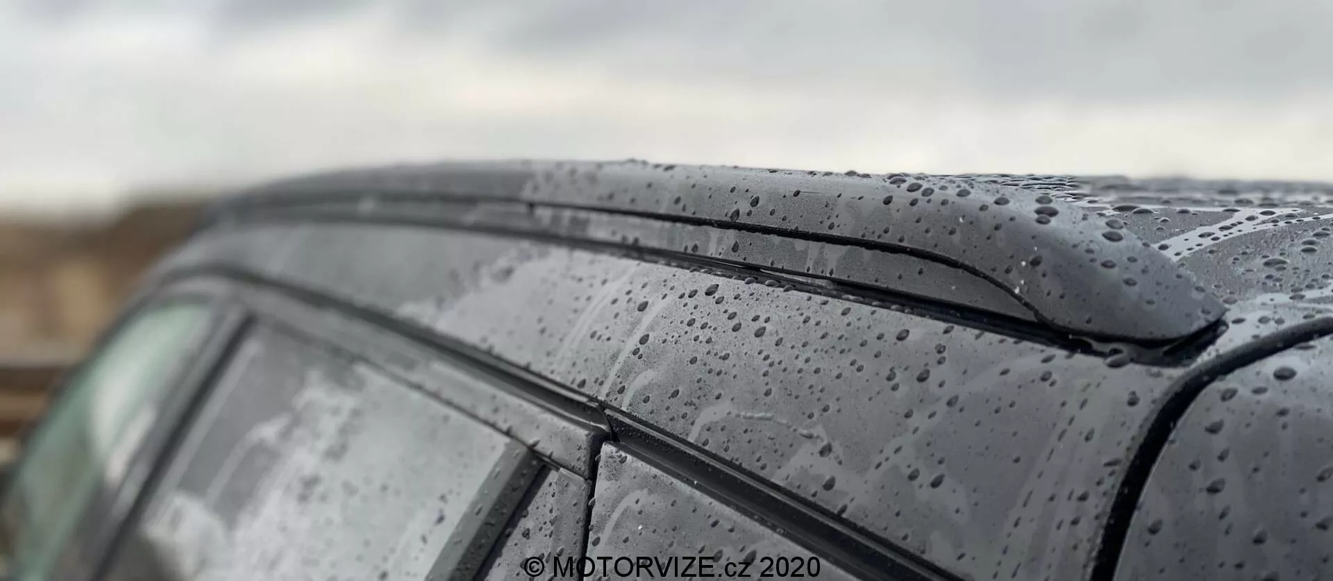Primer plano del techo y la ventana lateral del Peugeot 2008 1.2 PureTech 100 Active modelo 2020, destacando las gotas de lluvia sobre la superficie elegante. La imagen captura la pintura oscura y brillante con las gotas de lluvia y el diseño minimalista del borde superior de la ventana lateral. La poca profundidad de campo enfatiza la textura exterior del coche mientras desenfoca el fondo y partes del techo, mostrando una representación artística del coche en condiciones húmedas.