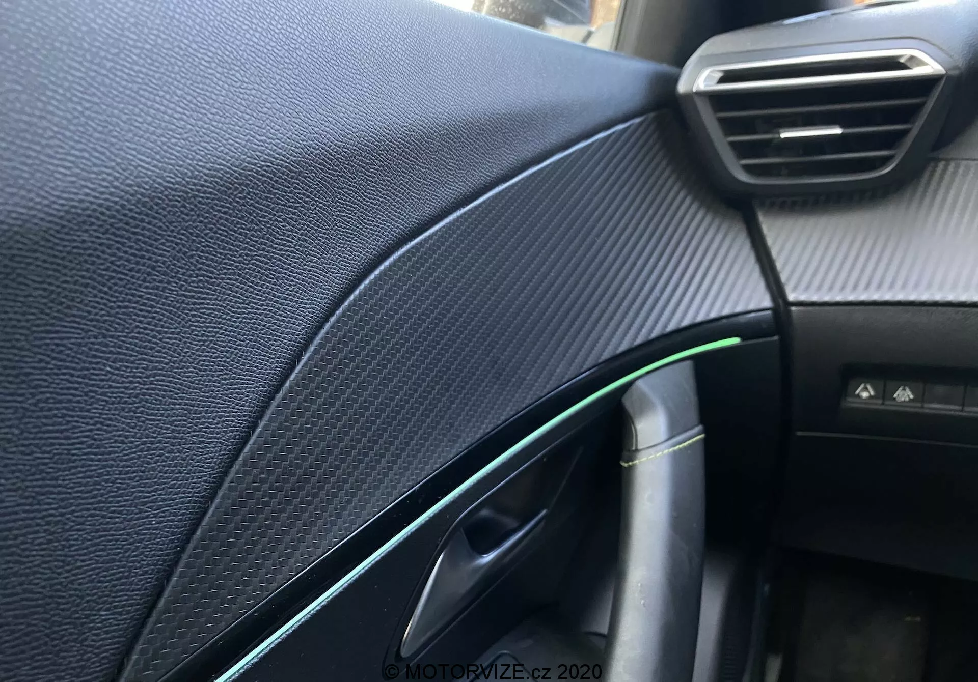 Vista detallada del tablero y detalles del lado del pasajero del Peugeot 2008 2020, centrada en el tablero texturizado negro sintético que imita a la fibra de carbono, la ventilación ajustable visible, la puerta parcialmente abierta con reflejo verde y un acento cromado, el tirador de la puerta, un cable con conector de carga, la luz de la cabina y un botón no identificado, capturada con poca profundidad de campo y luz natural.