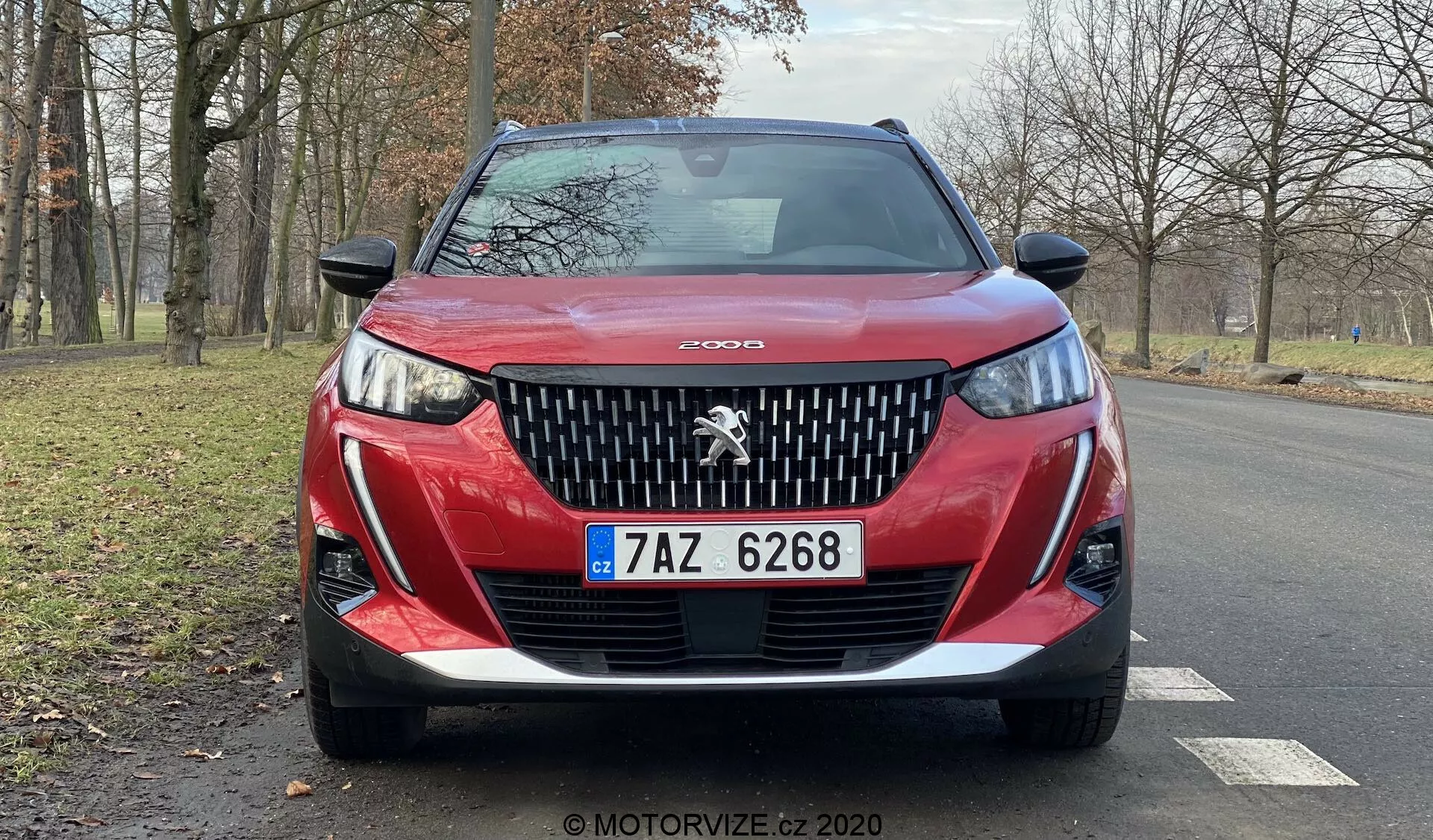 Vista frontal cercana de un Peugeot 2008 2020 rojo (Peugeot-2008-1-2-PureTech-100-Active) mostrando su detallada parrilla delantera con el prominente emblema del león de Peugeot, diseño agresivo de los faros con luces diurnas LED en forma de 'colmillos', parachoques inferior esculpido y placa de matrícula checa. El contorno del capó añade dinamismo y la imagen captura una vista parcial de las llantas de aleación y los espejos laterales en una carretera de asfalto seco con árboles sin hojas y una zona verde en el fondo, sugiriendo un entorno sereno y natural, posiblemente en otoño o invierno.