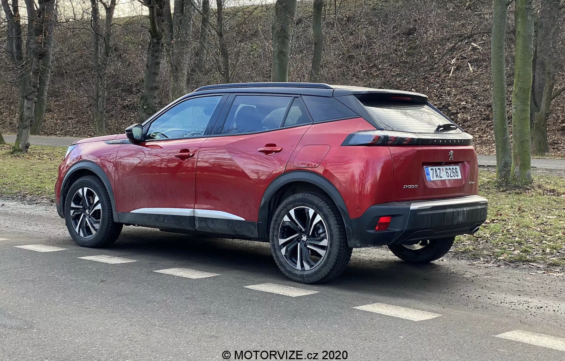 La imagen presenta un SUV Peugeot 2008 2020 de color rojo metálico brillante con un techo negro contrastante, ruedas de múltiples radios y neumáticos de perfil bajo. Fotografiado desde una vista trasera de tres cuartos, el SUV muestra sus líneas laterales marcadas, spoiler integrado, antena de aleta de tiburón, luces traseras LED horizontales estrechas y un parachoques trasero elegante con sensores de estacionamiento. El vehículo tiene manijas y espejos laterales del color de la carrocería con intermitentes integrados, ventanas traseras tintadas y una placa de matrícula central debajo de un posible lugar para una cámara de visión trasera.