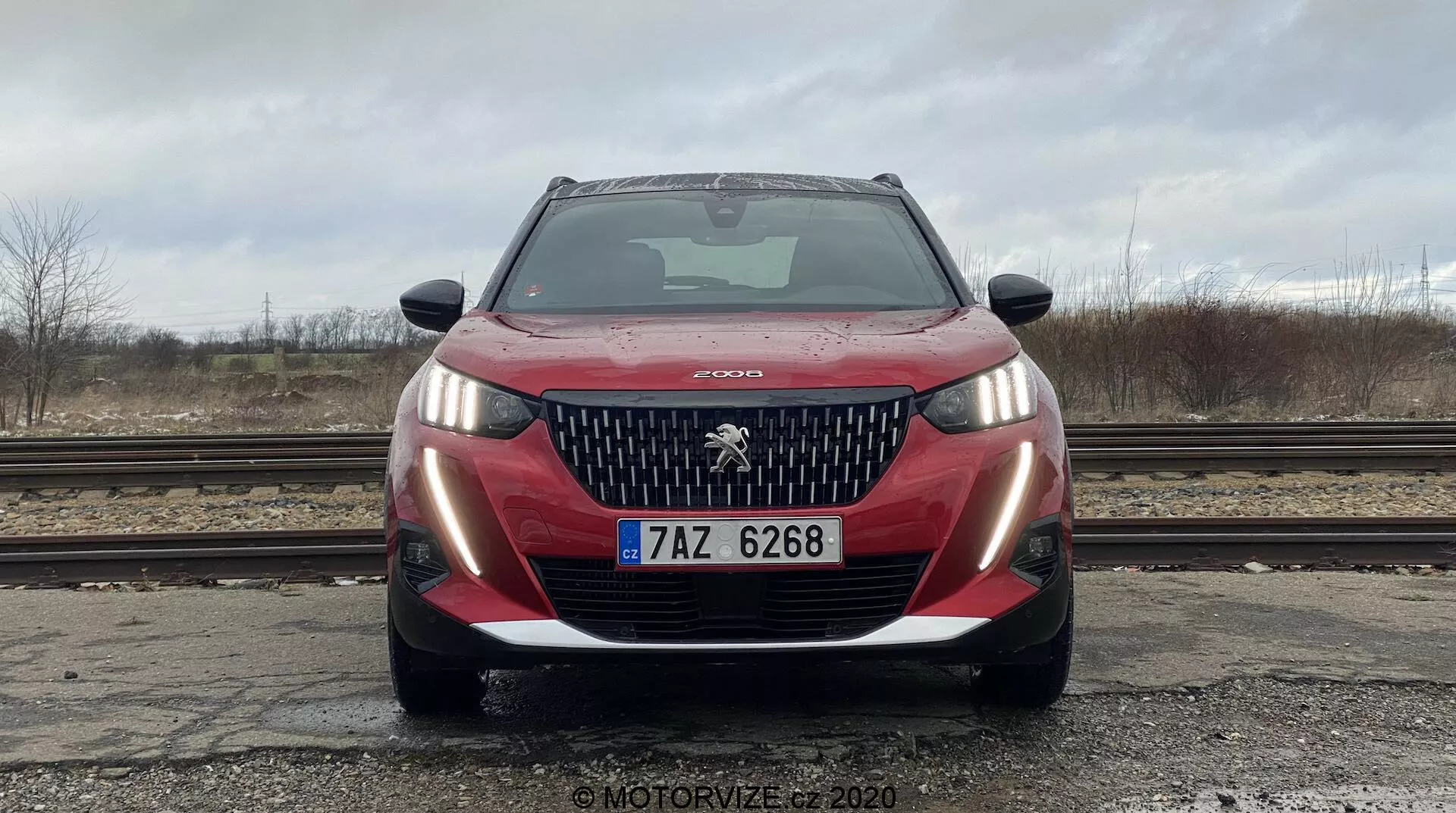 Vista frontal de un vehículo Peugeot 2008 2020 PureTech 100 Active rojo, fotografiado desde un ángulo bajo que destaca la parrilla delantera, el parachoques, los faros y el emblema de Peugeot. El coche está limpio con gotas de agua en el capó, parabrisas claro que refleja un cielo nublado. Las luces de estacionamiento están encendidas, la matrícula europea es visible. El escenario incluye suelo mojado, vías de tren, árboles desnudos y cielos grises, proporcionando una atmósfera industrial.