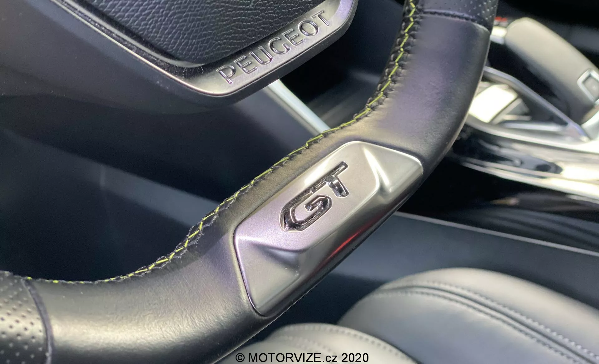 Primer plano del interior del Peugeot 2008 2020, destacando la insignia 'GT' en el volante de cuero perforado con costuras verdes, subrayando deportividad y lujo. La imagen se centra en los detalles de la artesanía y el fondo desenfocado revela materiales interiores premium y un diseño moderno.