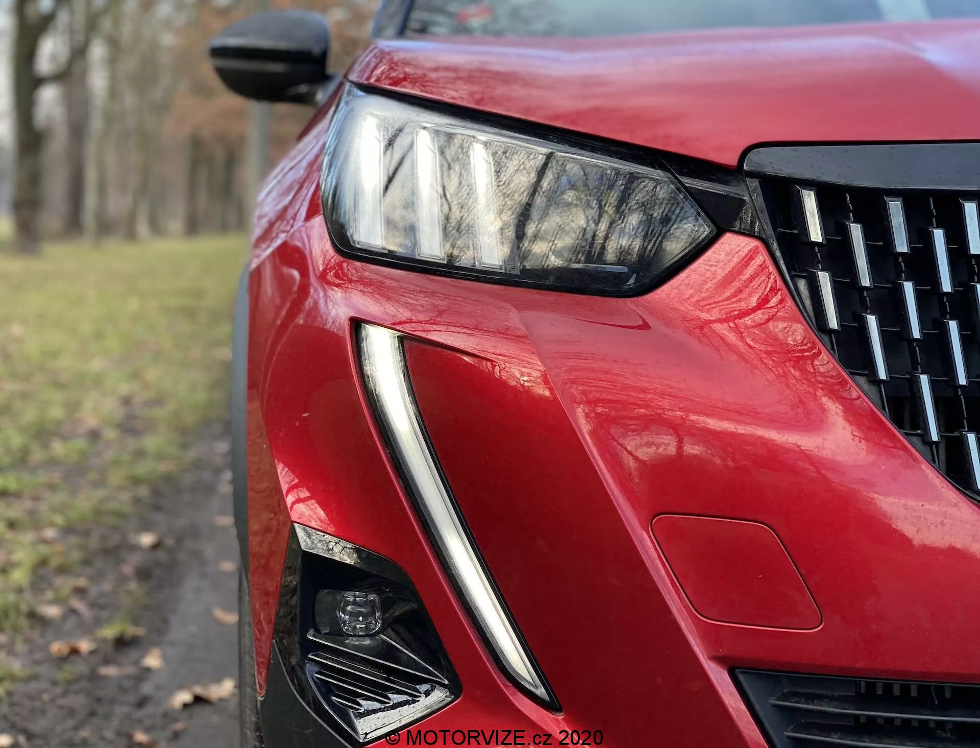 Vista en primer plano y ángulo bajo de la sección delantera izquierda del Peugeot 2008 2020 con acabado rojo brillante. La imagen resalta el elegante faro LED angular, el diseño afilado de la toma de aire y las líneas esculpidas, reflejando tecnología avanzada de iluminación y estética aerodinámica.