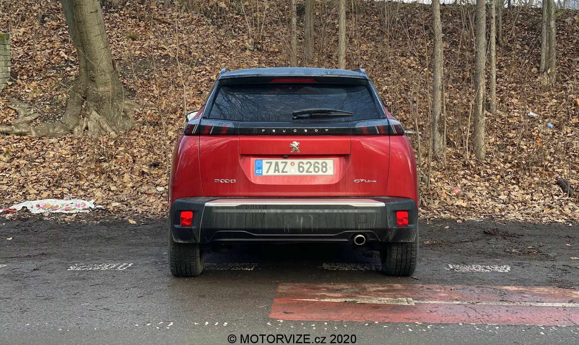 Vista trasera de un coche rojo estacionado al aire libre contra un fondo de matorrales o árboles sin hojas que sugiere otoño o invierno. Las características del coche incluyen un logotipo central y una placa de identificación en la tapa del maletero, matrícula, limpiaparabrisas trasero, alerón trasero, luces traseras modernas, reflectores y sensores de estacionamiento traseros en el parachoques, parachoques trasero prominente con un tubo de escape visible, placa de deslizamiento trasera, antena de aleta de tiburón en el techo y una pegatina despegada en el lado izquierdo inferior de la tapa del maletero.