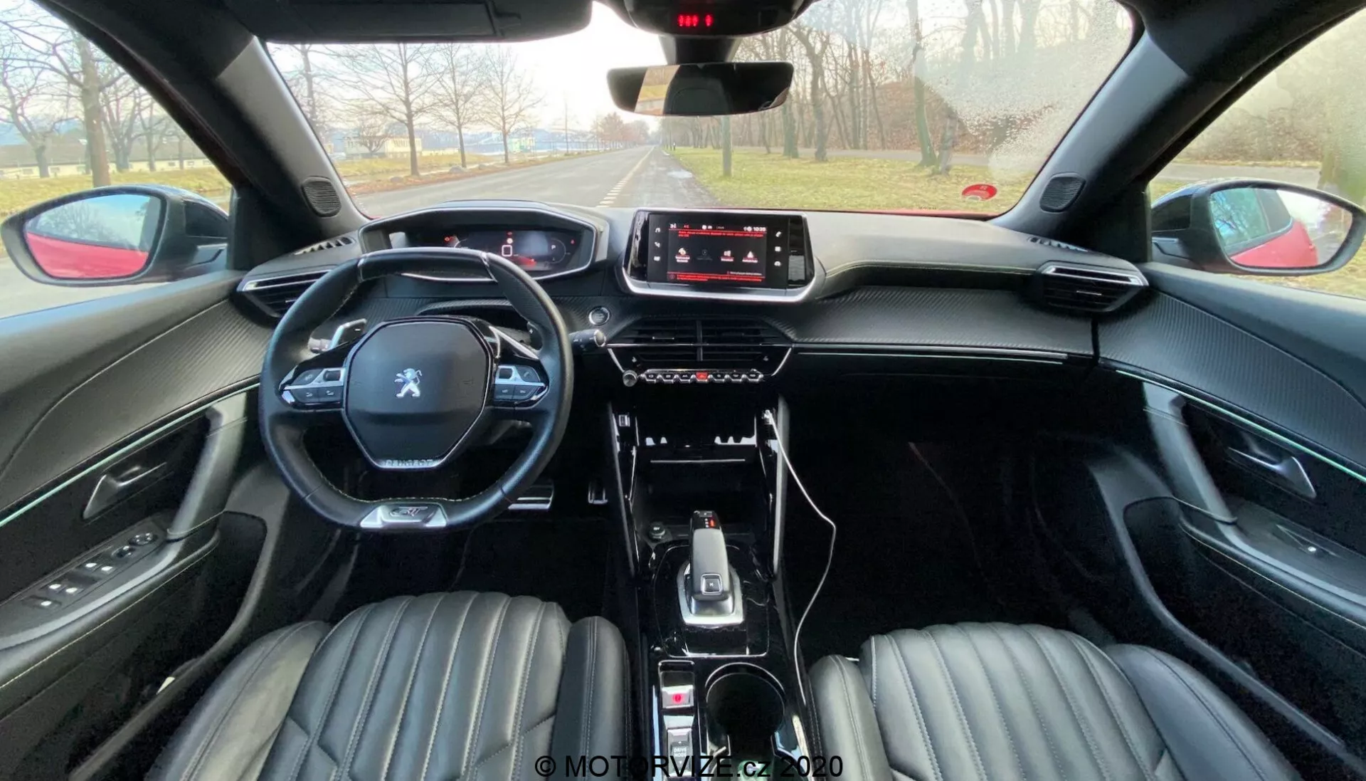 Vista interior del Peugeot 2008 2020 (peugeot-2008-1-2-puretech-100-active) desde la perspectiva del asiento trasero. La imagen muestra la fila delantera de asientos tapizados en cuero negro con diseño acanalado, reposacabezas integrados, un volante multifunción con el logotipo de Peugeot y una consola central moderna. El salpicadero cuenta con una pantalla táctil de infoentretenimiento con controles físicos mínimos, indicando un enfoque en la interacción táctil. El coche incluye una palanca de cambios automática, selectores de modos de conducción, freno de mano electrónico y compartimentos de almacenamiento. La atmósfera sugiere un día claro con una vista despejada de la carretera adelante.