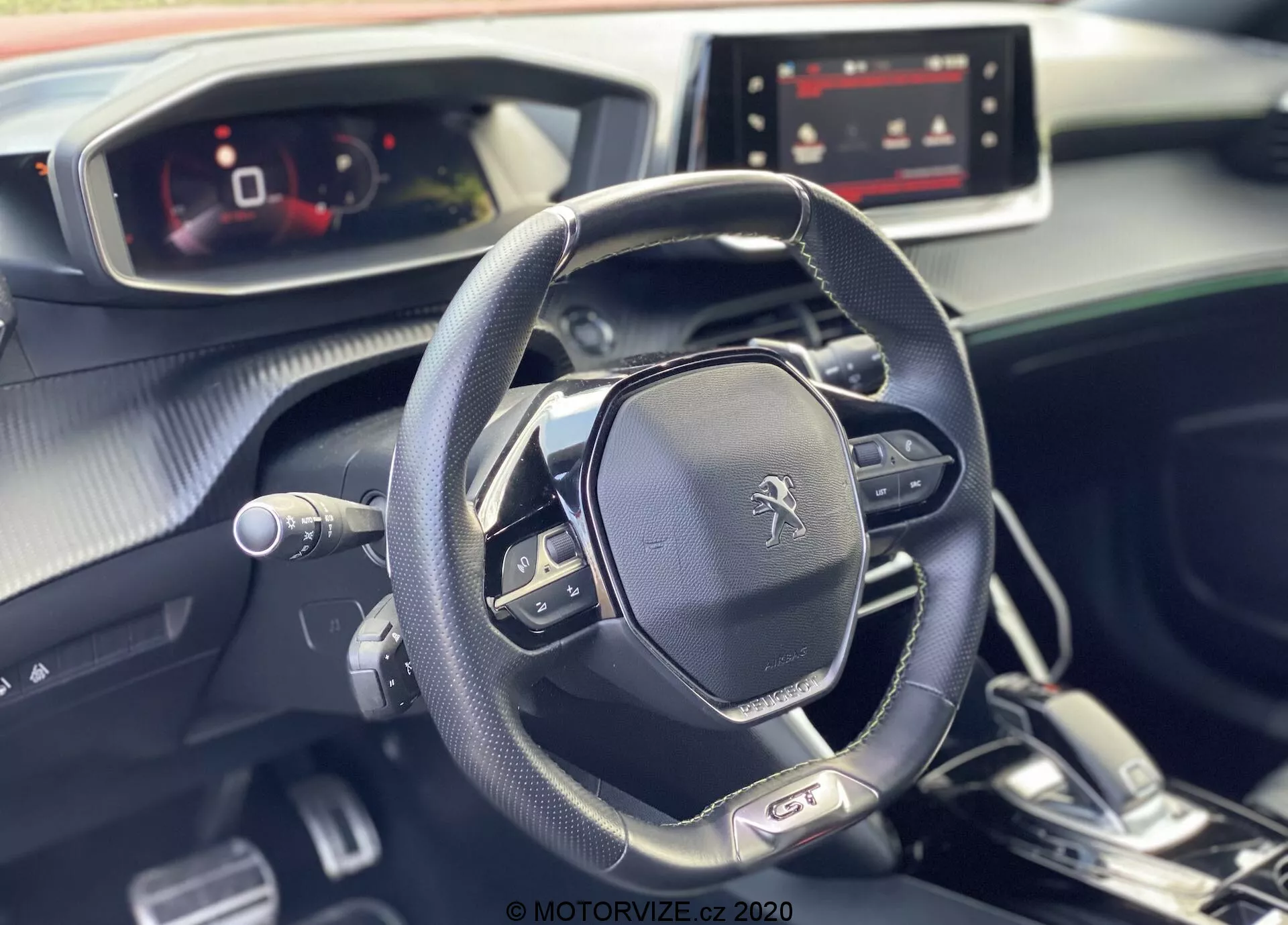 La imagen muestra el interior moderno y elegante del Peugeot 2008 2020, destacando el volante multifuncional y el panel de instrumentos totalmente digital desde la perspectiva del lado del pasajero. El emblema de Peugeot se muestra prominentemente en el volante, que cuenta con controles de medios, configuraciones de control de crucero y paletas de cambio, lo que sugiere una transmisión automática con opciones de cambio manual. El tablero de instrumentos, diseñado con una mezcla de materiales suaves al tacto y acentos metálicos, incluye una pantalla de infoentretenimiento encendida, que sugiere varias opciones de conectividad y navegación. La luz natural realza la atmósfera impecable y agradable del interior del coche.