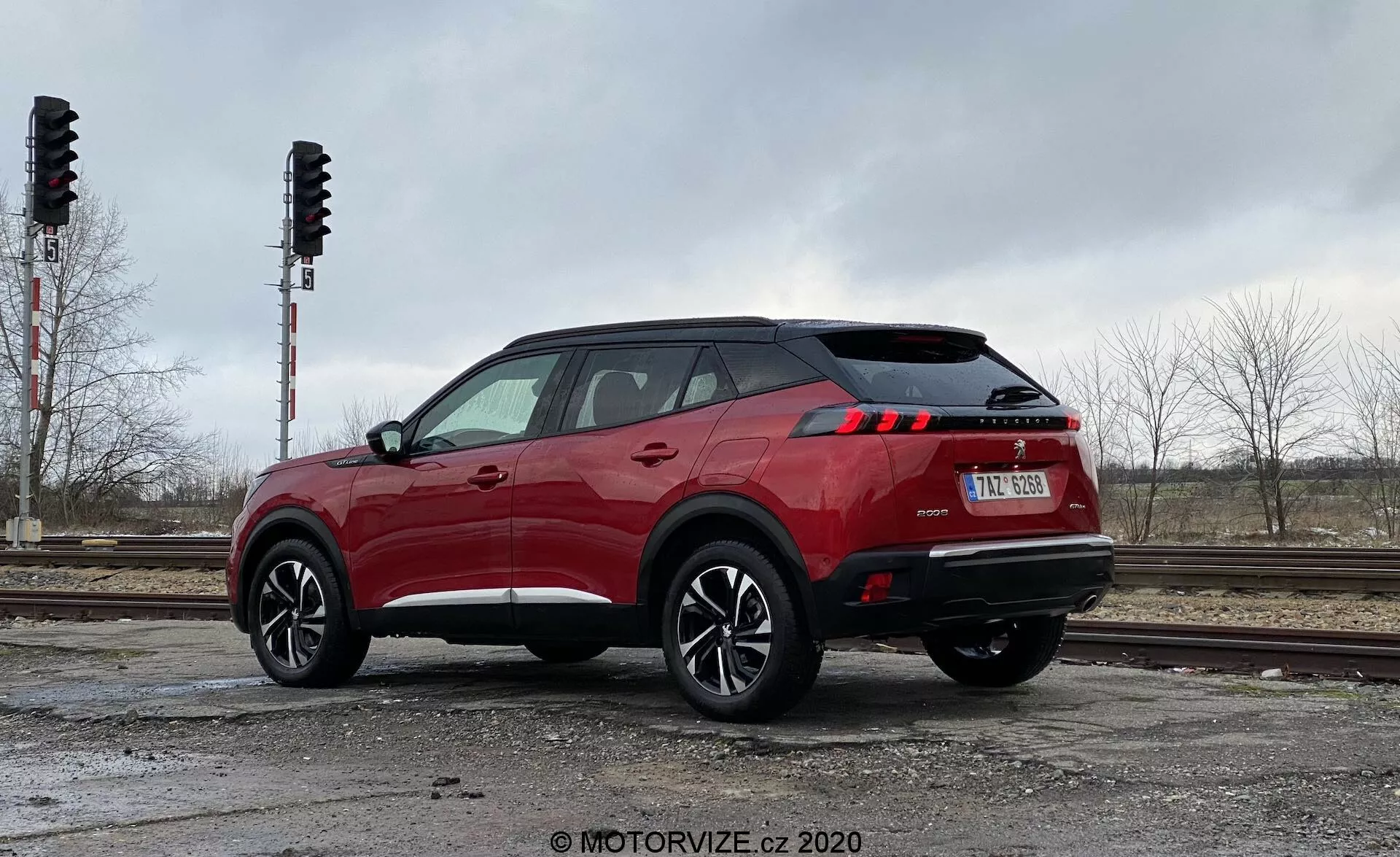 La imagen presenta un SUV moderno, específicamente el Peugeot 2008 2020 (1.2 PureTech 100 Active), capturado desde una vista trasera de tres cuartos. El ángulo ofrece una perspectiva clara del perfil lateral y trasero del vehículo, resaltando su diseño y dimensiones. Posicionado a la izquierda con espacio amplio a la derecha, el coche parece listo para moverse. La toma se realiza desde un ángulo bajo, cerca del suelo, ligeramente hacia arriba, acentuando la altura y posición del vehículo. La rueda trasera y el guardabarros son prominentes, mostrando el diseño de la rueda y la altura de la suspensión. El color rojo brillante de la carrocería contrasta con el fondo gris. La luz difusa de un día nublado proporciona visibilidad clara de los detalles, incluidas las luces traseras LED. El entorno incluye vías ferroviarias y árboles, indicando un ambiente urbano o industrial.