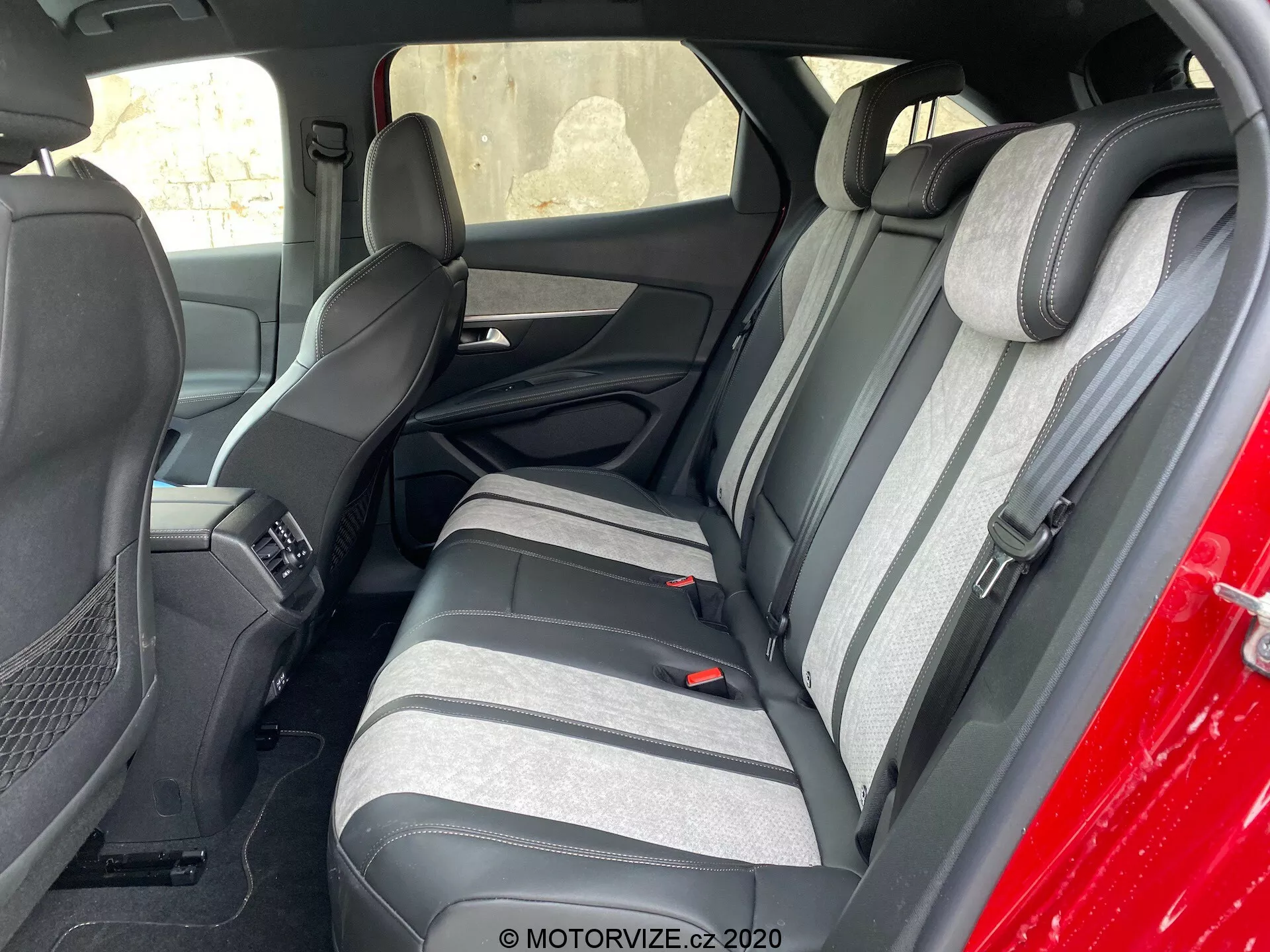 Toma interior de un Peugeot 3008 de 2021 enfocándose en la zona de los asientos traseros desde fuera, en ángulo hacia el lado del conductor. Los asientos traseros presentan una tela de dos tonos con grises oscuros y claros y un patrón de rayas texturizado. Los reposacabezas levantados a juego son visibles. Parte del asiento delantero y la consola central también están a la vista. Los asientos traseros tienen cinturones de seguridad de tres puntos y un reposabrazos con portavasos integrados. Los paneles de las puertas coinciden con los colores de los asientos e incluyen rejillas para altavoces y secciones de control de energía.