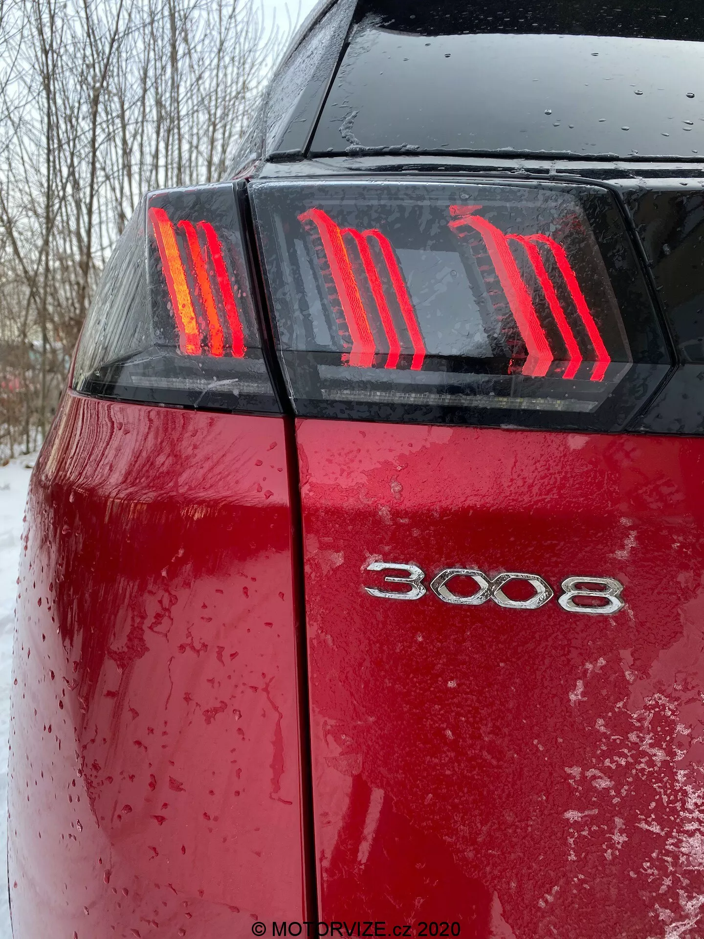 Acercamiento de la parte trasera del Peugeot 3008 2021 rojo desde un ángulo bajo, mostrando las luces traseras LED con tres franjas verticales en forma de garra y el distintivo del modelo '3008'. La superficie húmeda del coche, cubierta de gotas de agua y suciedad o nieve, resalta el uso reciente en condiciones meteorológicas adversas con condiciones de iluminación nublada.