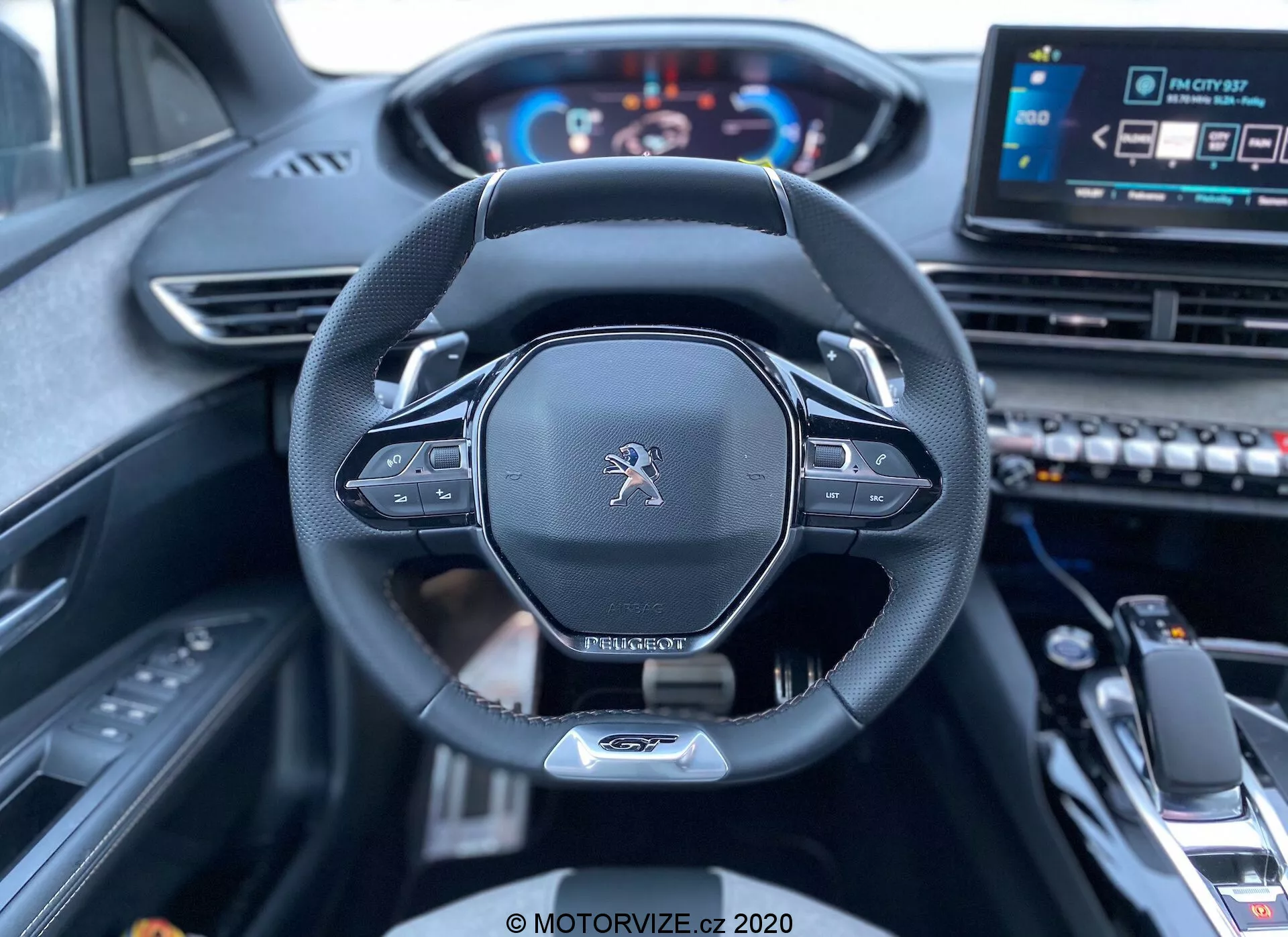 Vista interior desde el asiento del conductor de un Peugeot 3008 2021 mostrando el volante y el salpicadero. La imagen se centra en el volante enmarcado centralmente, con un logotipo de Peugeot, diseño moderno con parte inferior plana, borde de cuero y controles montados probablemente para ajustes de audio y crucero. Detrás del volante son visibles las paletas para cambio manual. Un cuadro de instrumentos digital muestra el velocímetro y el tacómetro iluminados, indicando que el vehículo está encendido. A la derecha, un sistema de infoentretenimiento con pantalla táctil y varios botones de control son parcialmente visibles junto con el selector de marchas y reposabrazos central en el primer plano inferior, sugiriendo un vehículo moderno y tecnológicamente avanzado con un interior de alta gama.