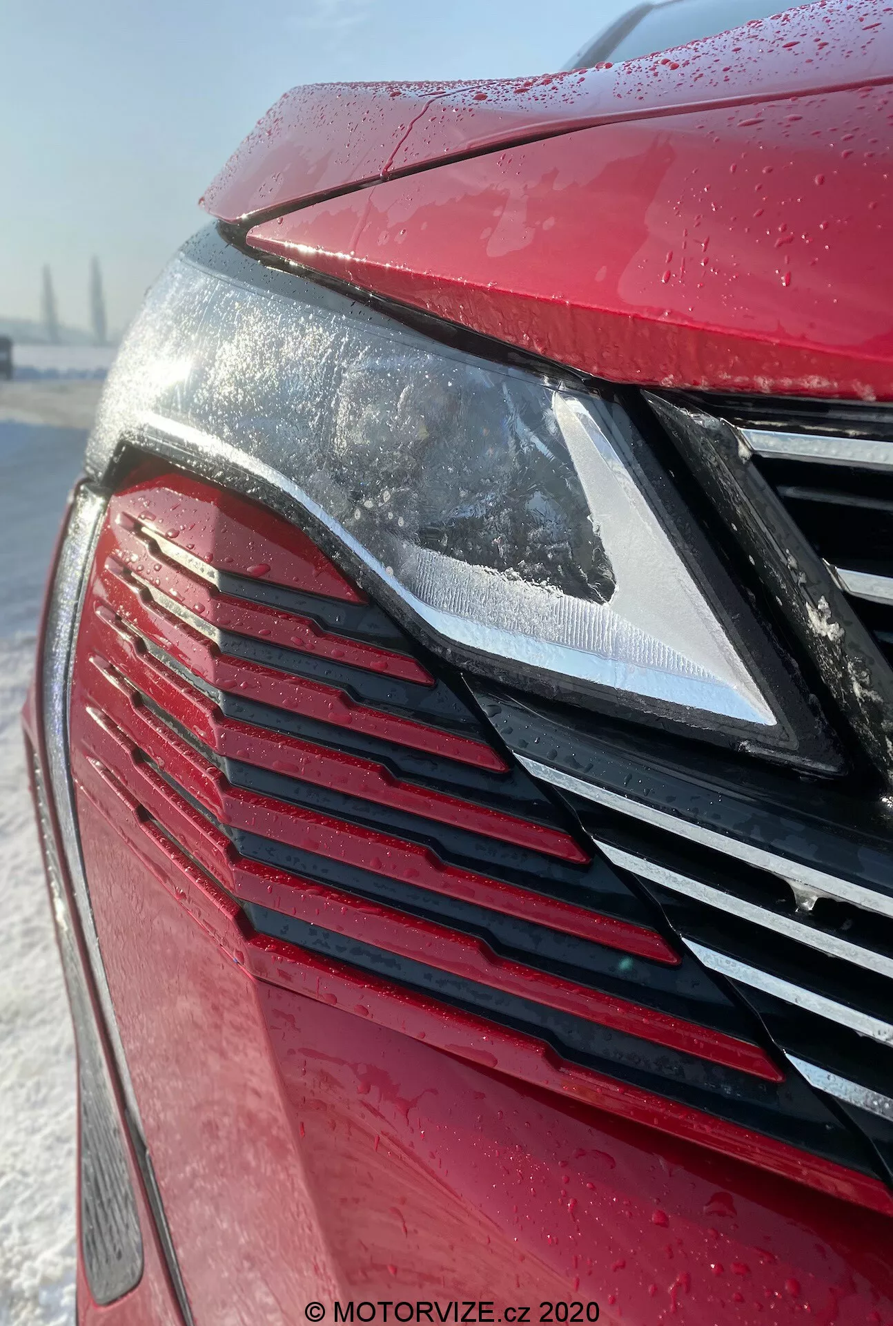 Vista detallada de la parte delantera del Peugeot 3008 2021, enfocándose en el faro LED moderno y la parrilla con lamas verticales en un exterior rojo metálico cubierto de gotas de agua, insinuando lluvia reciente o escarcha. El fondo nevado sugiere condiciones invernales o detallado de cuidado del automóvil.