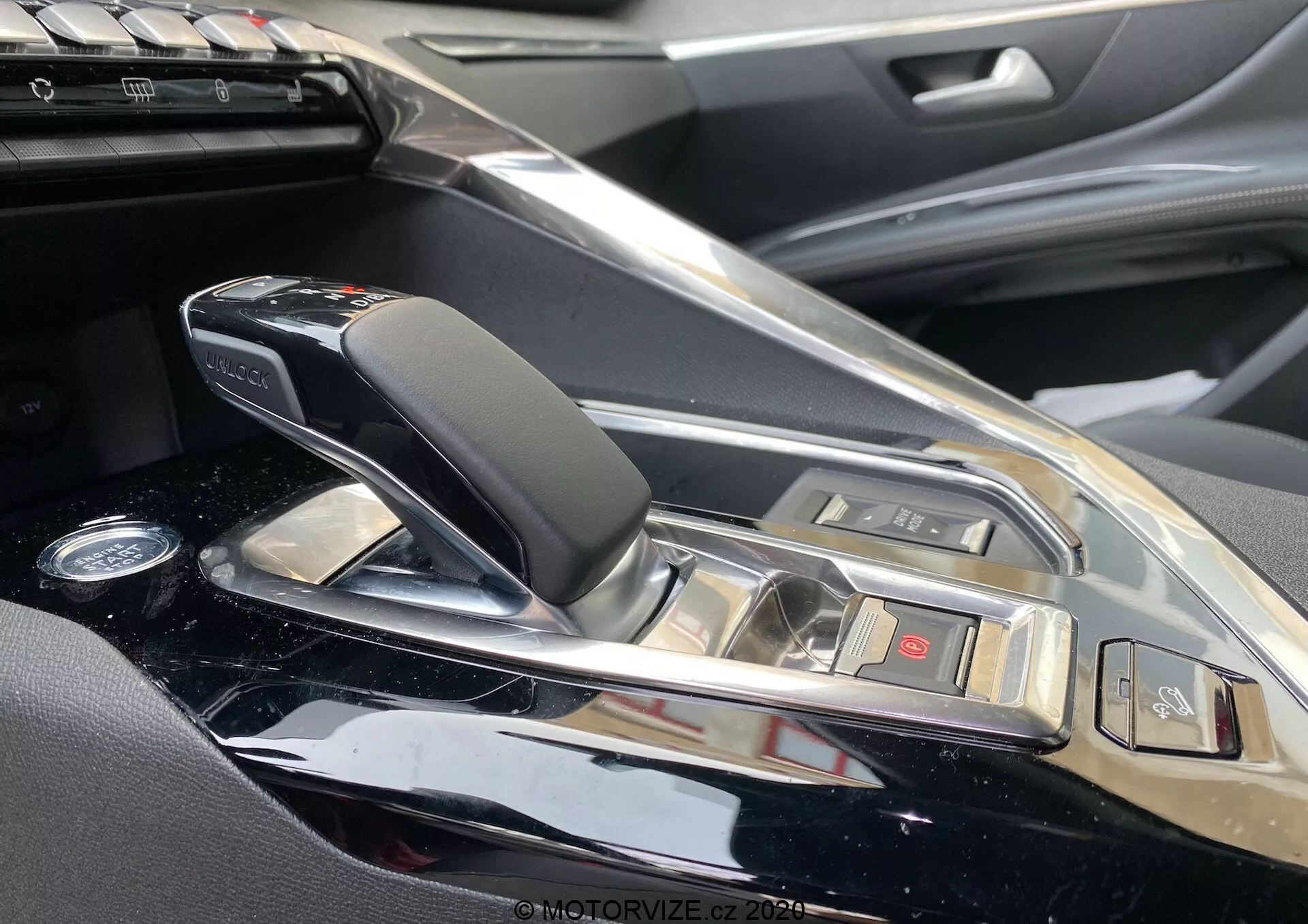 Vista de primer plano de la consola central y el área de la palanca de cambios del Peugeot 3008 2021 desde el lado del pasajero. Presenta una palanca de cambios automática moderna y elegante con detalles de cuero y cromo, rodeada de un acabado de consola brillante. Se ven los botones para varias funciones del vehículo y una vislumbre del sistema de infoentretenimiento, lo que indica un interior de coche premium con materiales de alta calidad y características tecnológicas sofisticadas.