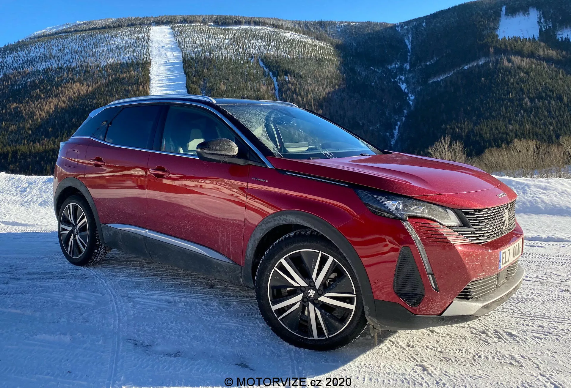Imagen de un coche moderno, probablemente un Peugeot 3008 2021, tomada desde una perspectiva de tres cuartos frontal. El ángulo muestra el diseño distintivo de los faros LED del coche, la parrilla con patrón de peine y el emblema en la parte delantera. El parachoques delantero muestra secciones de entrada de aire y alojamientos de luz antiniebla. El capó presenta líneas esculpidas, contribuyendo a la aerodinámica, mientras que el amplio parabrisas indica una buena visibilidad frontal. Las llantas de aleación multirradio sugieren un nivel de equipamiento más deportivo y superior. La línea de techo elegante y las líneas de la carrocería dinámicas insinúan un crossover o SUV. Los espejos laterales con indicadores de dirección integrados y molduras laterales o acentos visibles. El escenario es una carretera nevada con un fondo montañoso, lo que implica capacidad para diversos terrenos.