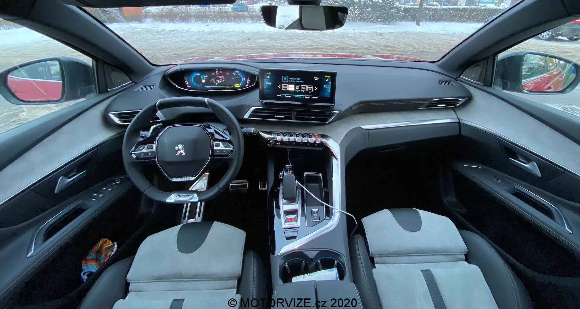 Vista interior del Peugeot 3008 2021, mostrando un volante multifuncional envuelto en cuero con controles montados, un panel de instrumentos digital, un gran sistema de infoentretenimiento con pantalla táctil en la consola central, un selector de marchas electrónico, asientos delanteros de diseño bicolor y una mezcla de materiales prácticos y lujosos. La foto está tomada desde el asiento trasero, proporcionando una vista amplia de la cabina destacando el diseño y las características tecnológicas.